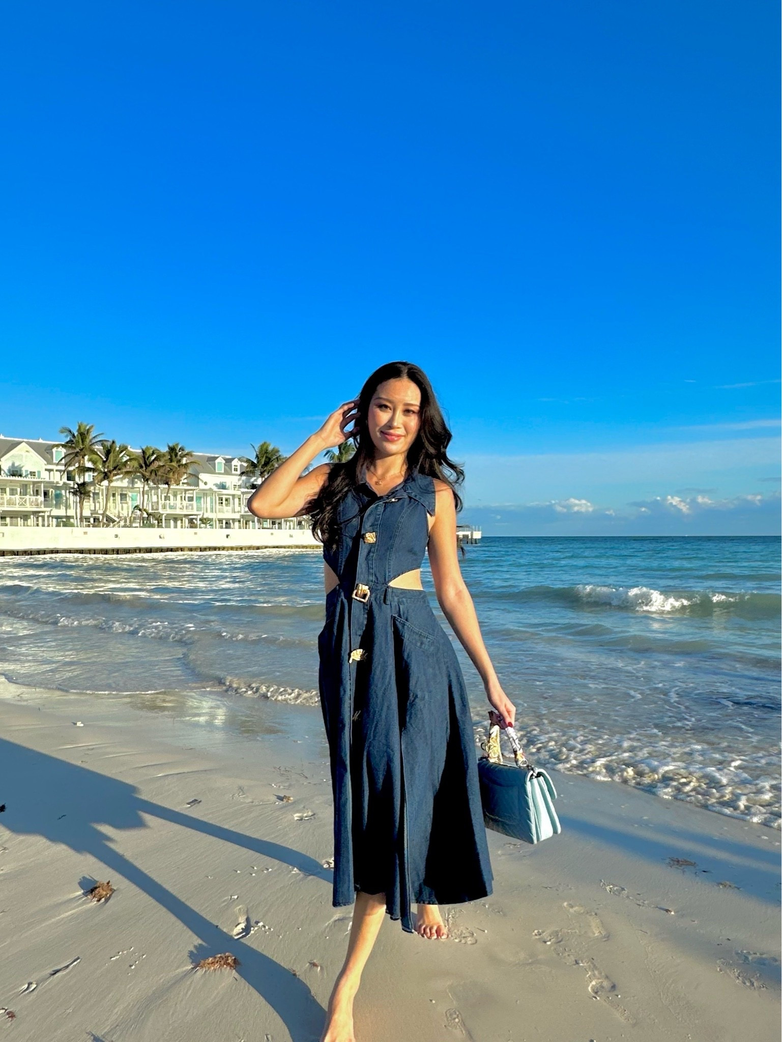 a dark denim sleeveless midi dress with side cutouts and gold buckle hardware, styled with a structured powder blue top-handle bag. the denim fabric dresses up or down effortlessly and the cutout detailing keeps it feeling fresh and modern. perfect for beach town days, resort brunches, summer date nights, and vacation strolls. shop this look linked above.