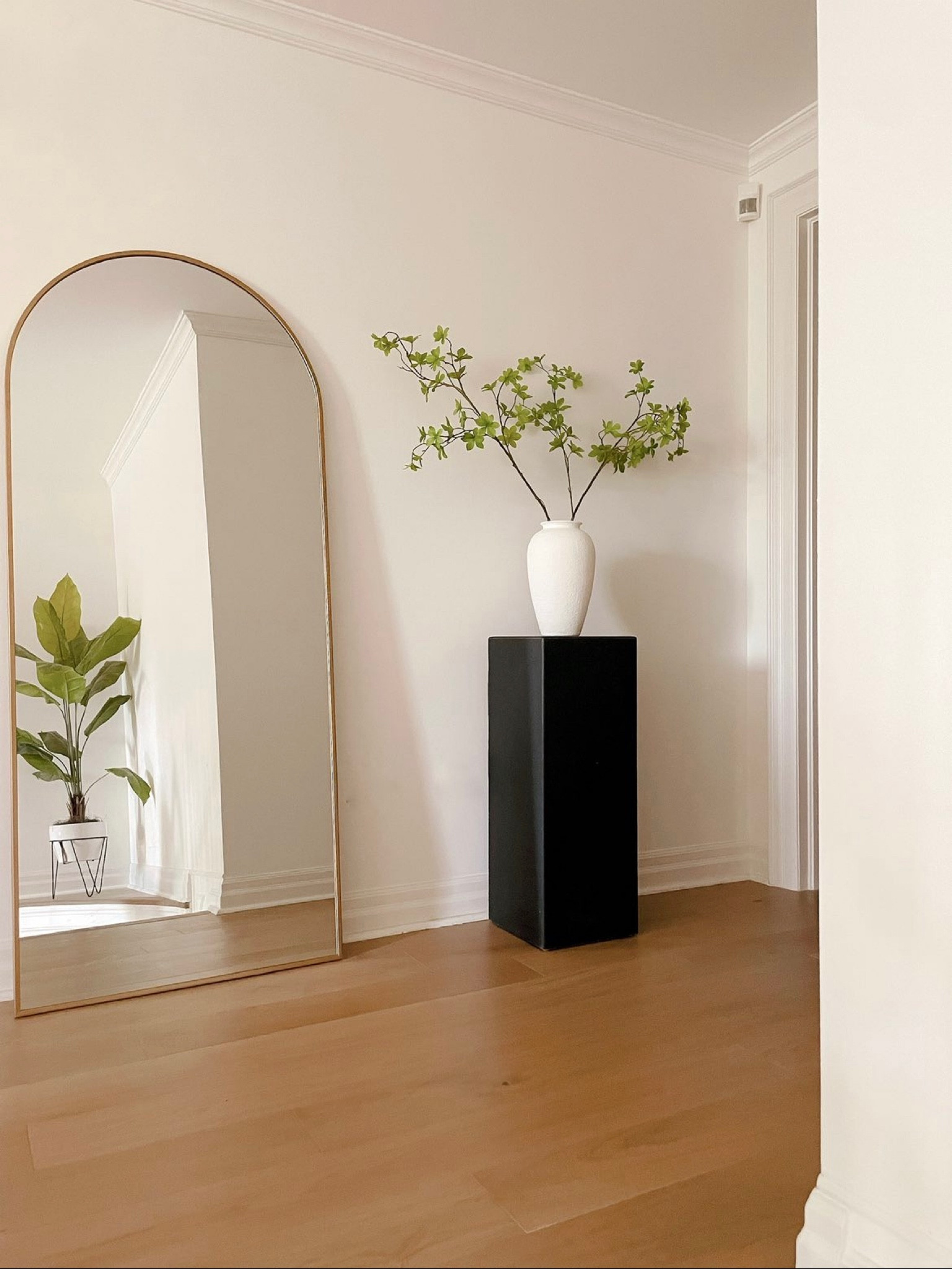 A simple home entrance design with a tall black art display pedestal holding a simple white vase and long faux maple stems and a gold arched floor mirror

#LTKunder100 #LTKFind #LTKhome