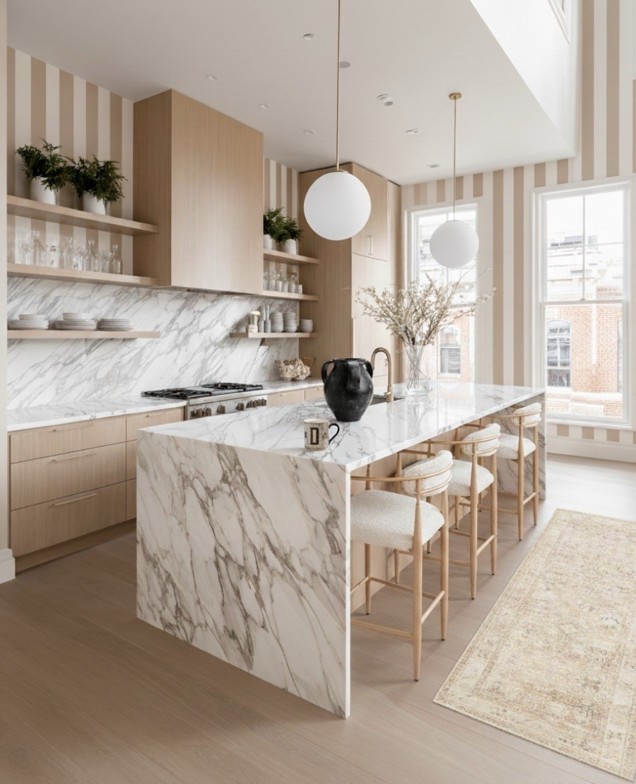 A timeless kitchen that redefines modern elegance.
This light-filled space pairs soft oak cabinetry with Calacatta marble countertops and backsplash, creating a luxurious yet inviting atmosphere. The striped taupe-and-cream wallpaper adds subtle pattern and visual height, while matte brass accents and minimalist globe pendants introduce a refined warmth.

Open shelving keeps the look airy, displaying curated glassware and ceramics that complement the kitchen’s soft neutral palette. The textured boucle bar stools and vintage rug add a cozy layer of character to an otherwise sleek, architectural space — proving that calm and personality can coexist beautifully. 

#LTKFindsUnder100 #LTKHome #LTKSaleAlert