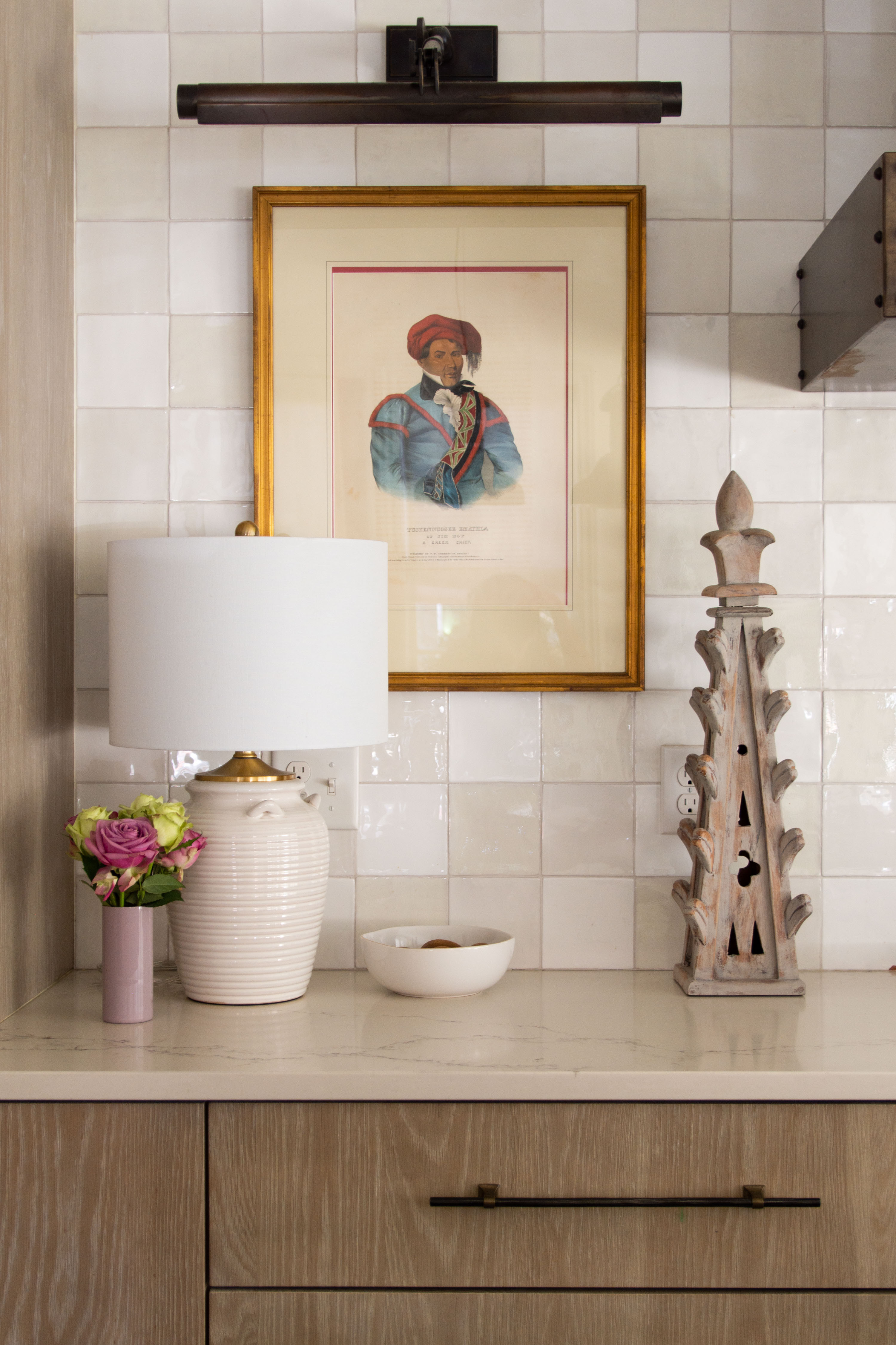 Warm and inviting kitchen corner styling inspiration featuring a mix of classic and modern design elements. The white ceramic table lamp with a linen shade adds soft lighting, while fresh roses in a blush pink vase bring a pop of color. A gold-framed vintage portrait hangs against a neutral zellige tile backsplash, highlighted by a sleek bronze picture light. The marble-look quartz countertop is styled with a simple white bowl and a rustic carved wood finial accent for added texture. Perfect for transitional kitchen decor, countertop styling ideas, and timeless interior design inspiration. 

 