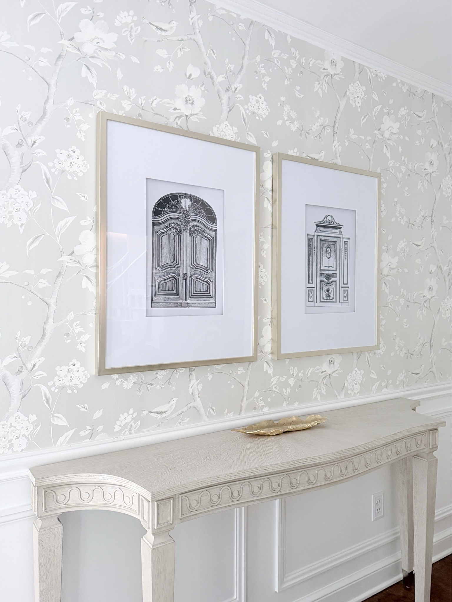 Gorgeous silver metallic luster Chinoiserie neutral wallpaper on a budget with oversized line drawing French door prints in champagne gold over a neoclassical console table with a gold decorative feather tray