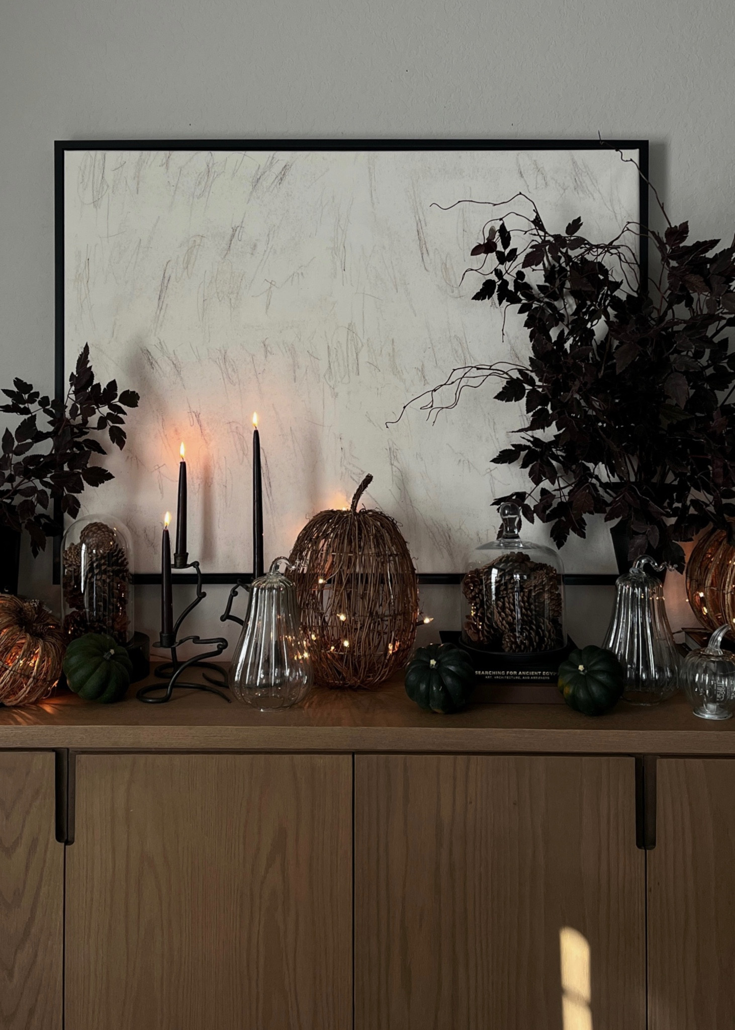 A Moody yet modern autumn and fall sideboard styling idea. Dark purple, so leaves and branches mixed with brown stems, wicker, pumpkins, glass, pumpkins, wavy candleholders, pinecones under glass against a hand drawin squiggly artwork