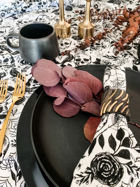Halloween place setting with black and white skull tablecloth and matching napkins, purple, dried eucalyptus, black dinnerware, gold, flatware, black cups, and gold candlestick holders. Get ready for your Halloween parties with this Halloween table decor.

#LTKHoliday #LTKhome #LTKHalloween