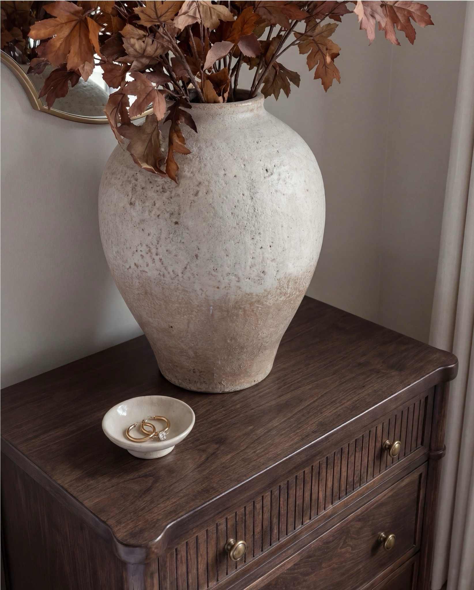 Shop the Look:
• White upholstered wingback bed frame
• White cotton duvet cover + pillow shams
• Textured throw pillows (rust + black patterned)
• Chunky knit throw blanket (warm brown)
• Vintage-style brown area rug
• Dark wood nightstands with brass knobs
• Ceramic table lamp (neutral glaze)
• Brass globe wall sconces
• Framed floral artwork (muted tones)
• Arched brass wall mirrors
• Upholstered bench at foot of bed (camel brown)
• Cream accent chair with wood arms
• Black fluted dresser with brass pulls
•Ceramic beige decor bowl
•Decor wood beads 
•Decor book
• Large ceramic floor table vase (earthy clay tone)
• Decorative wood bead garland
• Sheer white curtain panels
•Decor tray
•Hurricane candle holder
Wood Jewelery box 

#LTKHome #LTKSaleAlert