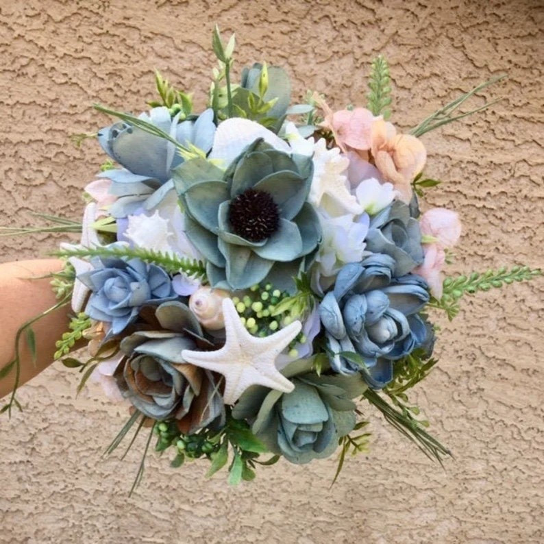 For the bride dreaming of ocean breezes and sandy shores, this beach-inspired bouquet by ForeverFlowersbyGigi is the perfect finishing touch. Handcrafted from eco-friendly sola wood flowers in soft blues, airy greens, and creamy ivory, it’s accented with natural seashells and finished with a burlap-wrapped handle for that relaxed coastal feel. Lightweight, sustainable, and designed to last long after your vows are exchanged, this bouquet gives you timeless beauty without the environmental impact of fresh florals. A stunning keepsake for seaside ceremonies, destination weddings, or any bride who loves a touch of the shore.

#LTKWedding #BeachBride #EcoFriendlyWedding #SolaWoodFlowers #CoastalWedding #SustainableBride #BridalBouquet #EtsyFinds

#LTKWedding #LTKHome #LTKSeasonal