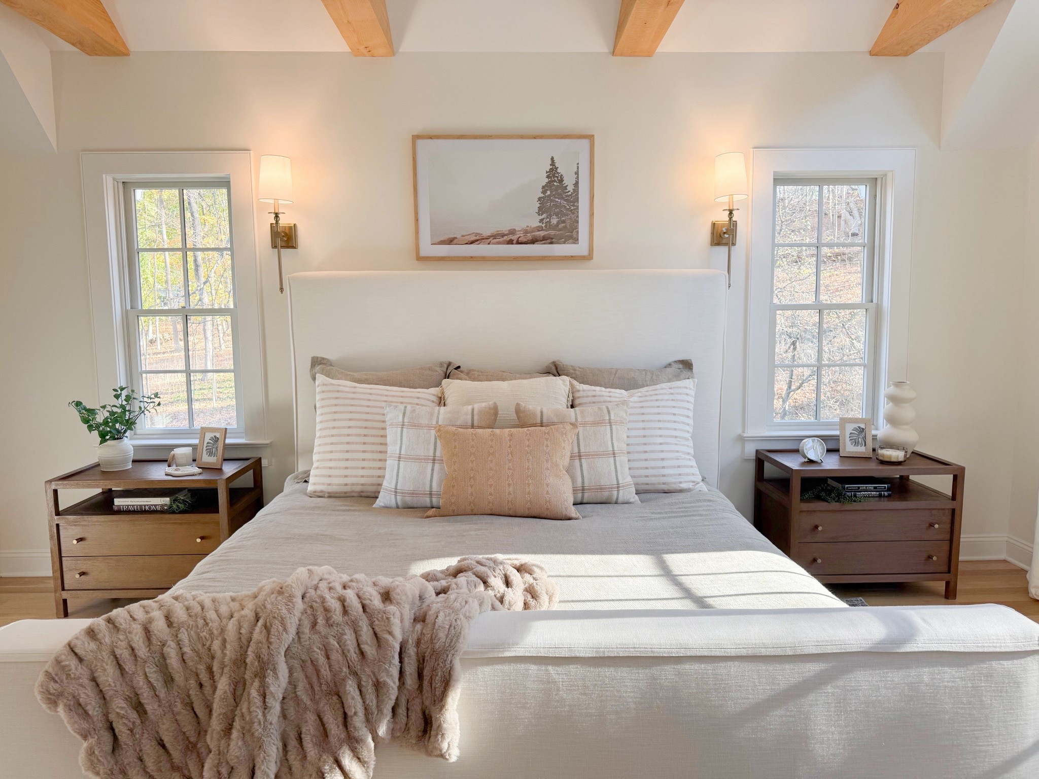 A cozy and inviting view of the Mount Kisco primary bedroom, showcasing a beautifully styled bed flanked by matching nightstands and elegant wall sconces. Layered in warm, earthy textures—from plush bedding and soft throws to natural wood accents—the space exudes comfort and timeless sophistication. A serene and thoughtfully designed retreat perfect for a good night sleep. 
