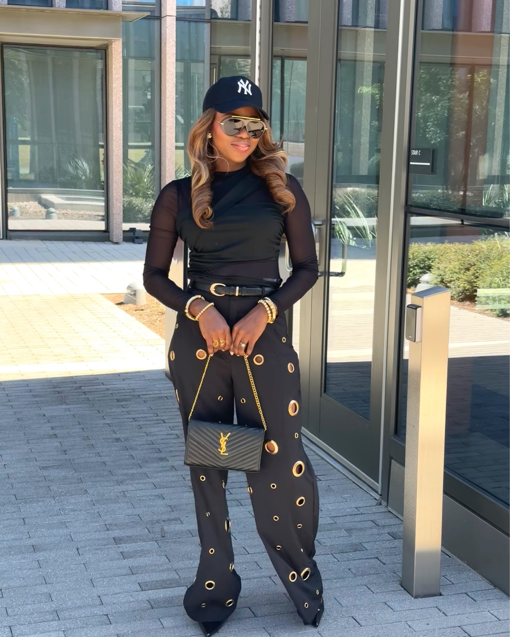 Elevated monochrome black outfit featuring a sheer long-sleeve top, high-waisted grommet statement pants with gold accents, luxury chain shoulder bag, NY baseball cap, oversized sunglasses, and sleek pointed heels for a chic modern street-style look.



#LTKU #LTKootd #LTKgrwm