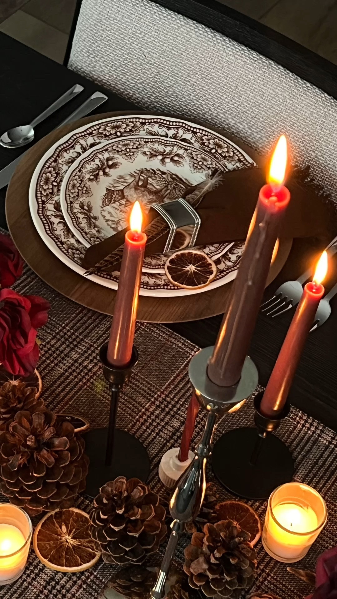 Ralph Lauren inspired tablescape, Ralph Lauren lodge aesthetic, moody fall table decor, fall hosting, autumn tablescape, plaid table runner, brown transferware dishes, stag plates, woodland dinnerware, wood chargers, chocolate brown cloth napkins, silver napkin rings, feather napkin accents, burgundy roses, silver centerpiece vase, brown stemmed glasses, silver flatware, taper candles, mixed height candle holders, votive candles, layered candlelight, pinecone table decor, dried orange slices, rustic elegant table setting, cozy candlelit dinner, Thanksgiving table inspiration, collected table style