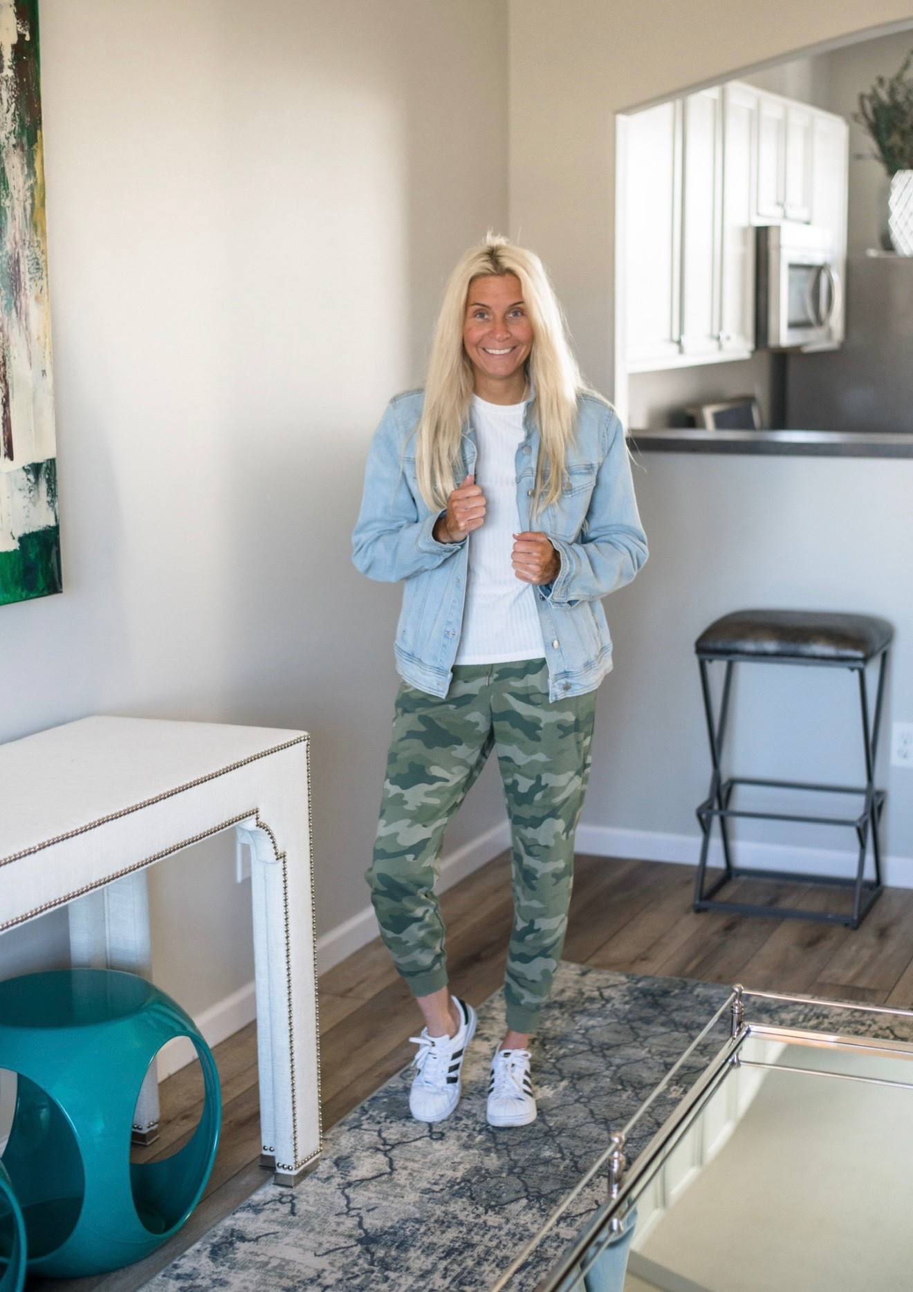 
A bright, lived-in space becomes the backdrop for a relaxed, effortlessly cool look. A blonde woman stands confidently in soft natural light, wearing a crisp white ribbed top layered under a light-wash denim jacket. Her long hair falls loosely around her shoulders, adding an easy, undone vibe. She pairs the top with tapered camo joggers that blend comfort and edge, finished with classic white sneakers for a laid-back, sporty feel.

The room’s neutral walls, abstract art, and textured blue-gray rug create a calm, modern environment that makes the outfit pop. The overall scene feels approachable and warm — the perfect mix of weekend comfort and everyday style.



#LTKdayinmylife #LTKHoliday #LTKGiftGuide