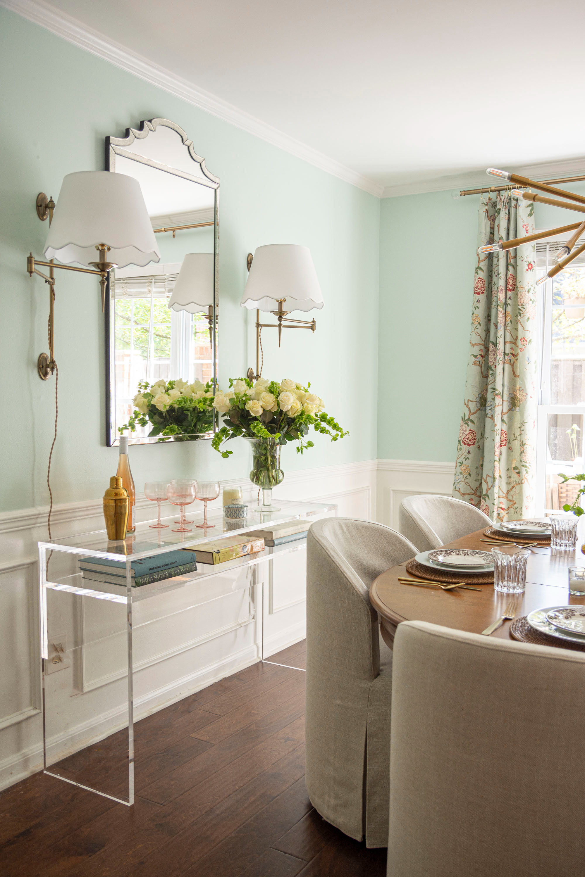 Light, bright, and timeless dining room inspiration. This elegant setup features a soft mint wall color paired with classic white wainscoting, a wood dining table, and upholstered slipcovered chairs. A gold and acrylic console table adds a touch of modern glam, styled with coffee table books, fresh florals, and barware essentials. The scalloped wall sconces and decorative mirror create a chic focal point, while floral patterned drapes bring in a cozy, traditional vibe. Perfect mix of modern, transitional, and classic dining room decor for an inviting yet sophisticated space. 

 