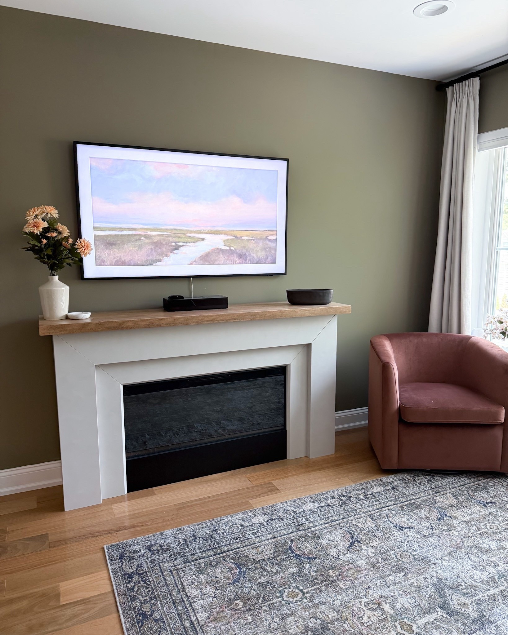 This cozy neutral living room features a Pottery Barn fireplace, Amazon vintage rug, and Target velvet swivel chair for a warm, layered look. The sage green wall color pairs beautifully with soft peach florals, a pleated lamp, and a matte black five-wick candle from Walmart. Styled with a Samsung Frame TV and textured wood mantel.

#LTKHome #LTKStyleTip #LTKOver40