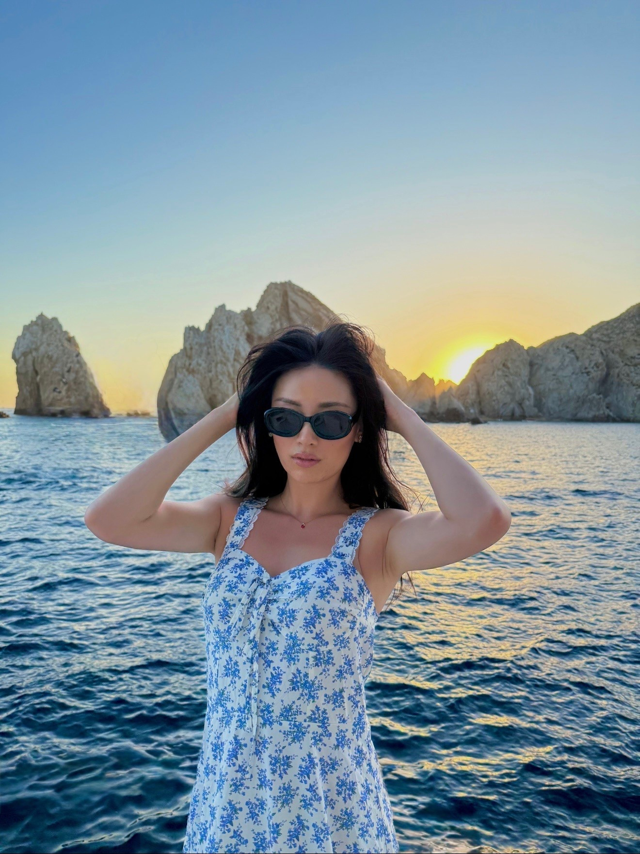 a white and blue floral sundress with lace trim straps and a sweetheart bow tie front, styled with black oval sunglasses for a golden hour sunset moment past the iconic land’s end rock formations in cabo san lucas. the romantic ditsy floral print and feminine silhouette make this the perfect resort sundress for sunset evenings. perfect for cabo vacations, baja resort getaways, sunset boat days, and mediterranean-inspired coastal trips. shop this look linked above.