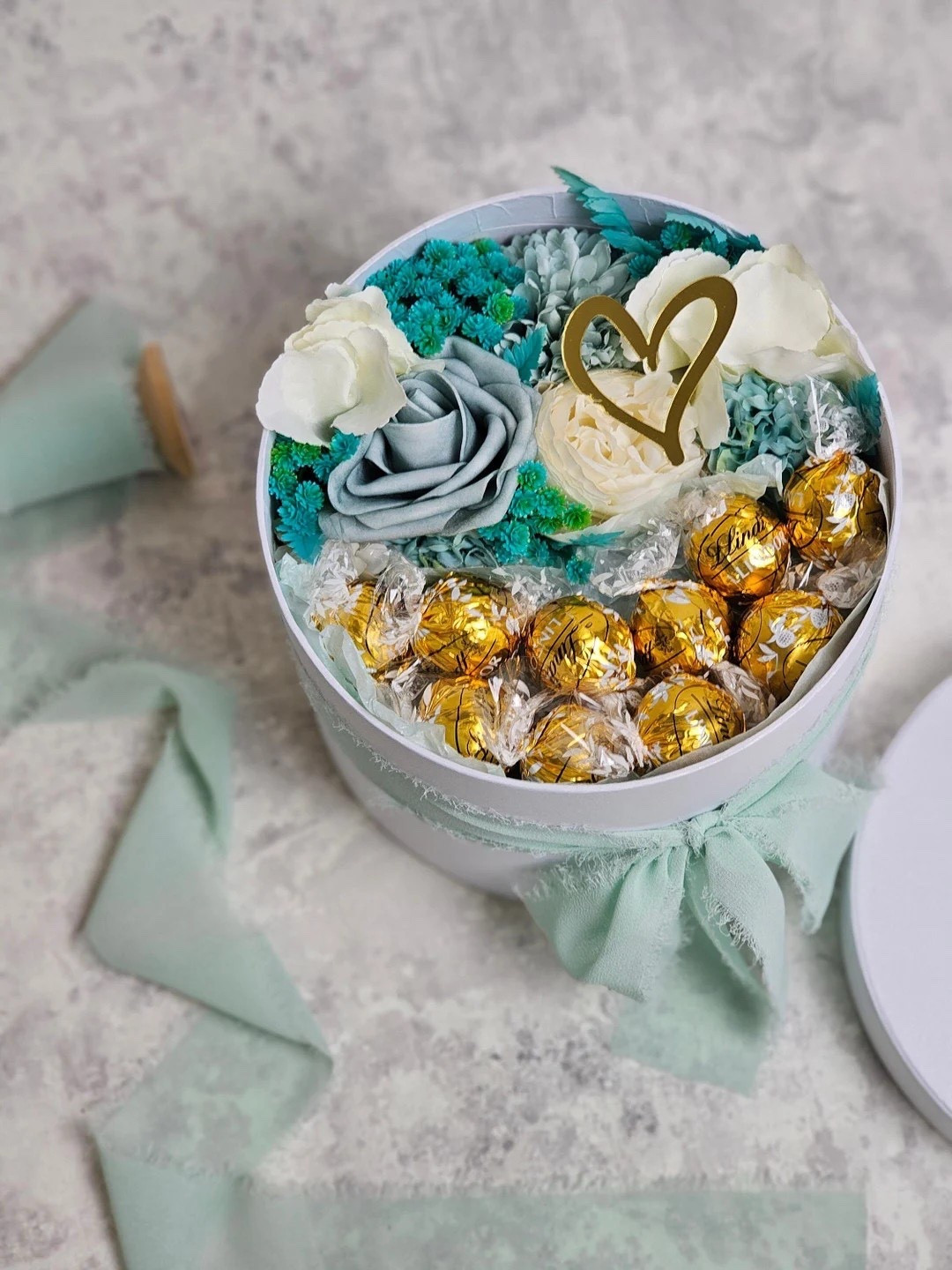 Elegant with a pop of color, this floral chocolate hat box by BlossomBeGifts is a gift that truly wows. Designed in soft white, blue, and turquoise tones, it features luxe silk and foam florals arranged on satin bedding, finished with chiffon ribbon and a gold heart ornament. Inside, decadent Lindt Lindor chocolates add the perfect sweet touch. Each box is handcrafted to order, making every arrangement a one-of-a-kind keepsake — beautifully packaged and ready to gift for birthdays, celebrations, or just because. 💙✨

#LTKgifts #GiftIdeas #LuxuryGifting #ChocolateGift #FlowerBox #HatBoxFlowers #SpecialOccasion #BlossomBeGifts #LTKhome

#LTKHome #LTKSeasonal #LTKFindsUnder50