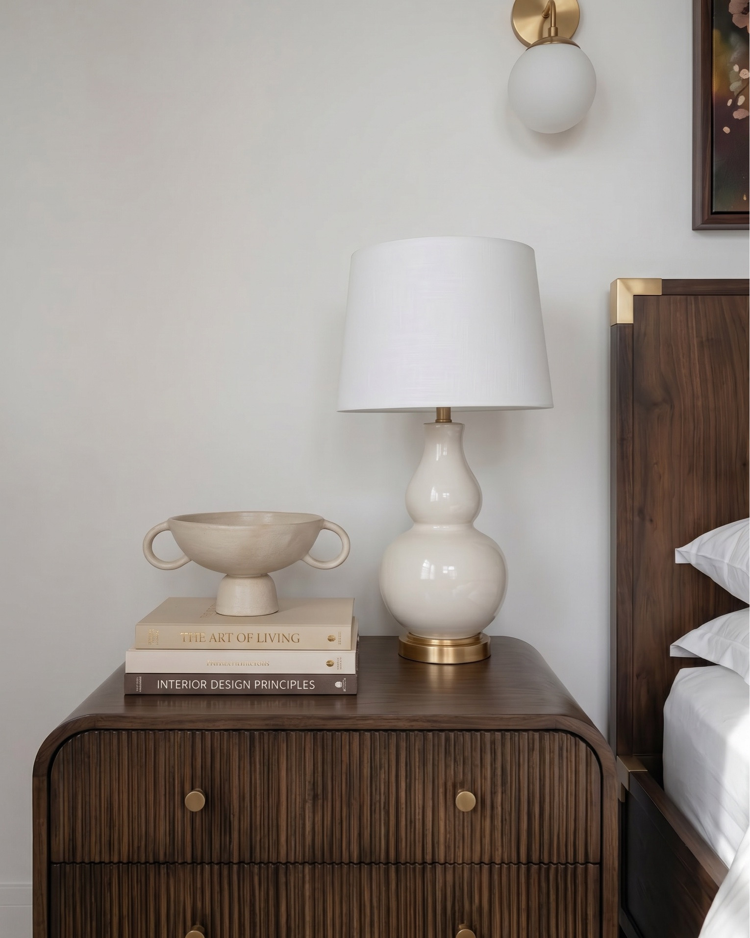 Warm modern bedroom design featuring a rich wood bed frame with brass accents, styled with crisp white bedding, textured neutral throw blanket, and terracotta accent pillows for a cozy, elevated look. A plush upholstered bench adds softness at the foot of the bed, layered over a geometric brown and cream area rug.
Matching wood nightstands with fluted detailing are styled with ceramic table lamps, decorative vases, and stacked books for a balanced, designer feel. Above the bed, a statement abstract art piece ties together warm earthy tones, complemented by modern brass wall sconces.
The space continues with a coordinating fluted wood dresser styled with neutral ceramic decor, a framed botanical print, a glass hurricane candle holder, and a decorative tray with small accessories. A cozy accent chair with soft upholstery and wood frame pairs perfectly with a matching round side table, topped with a candle for a warm, inviting touch.
 modern bedroom furniture, warm neutral bedroom decor, wood bed frame, upholstered bench, fluted dresser, designer nightstand styling, ceramic table lamps, abstract wall art, brass wall sconces, cozy accent chair, neutral home decor, earthy tone bedroom, luxury bedroom styling, high-end furniture look for less, transitional modern decor.

#LTKsale #LTKhome