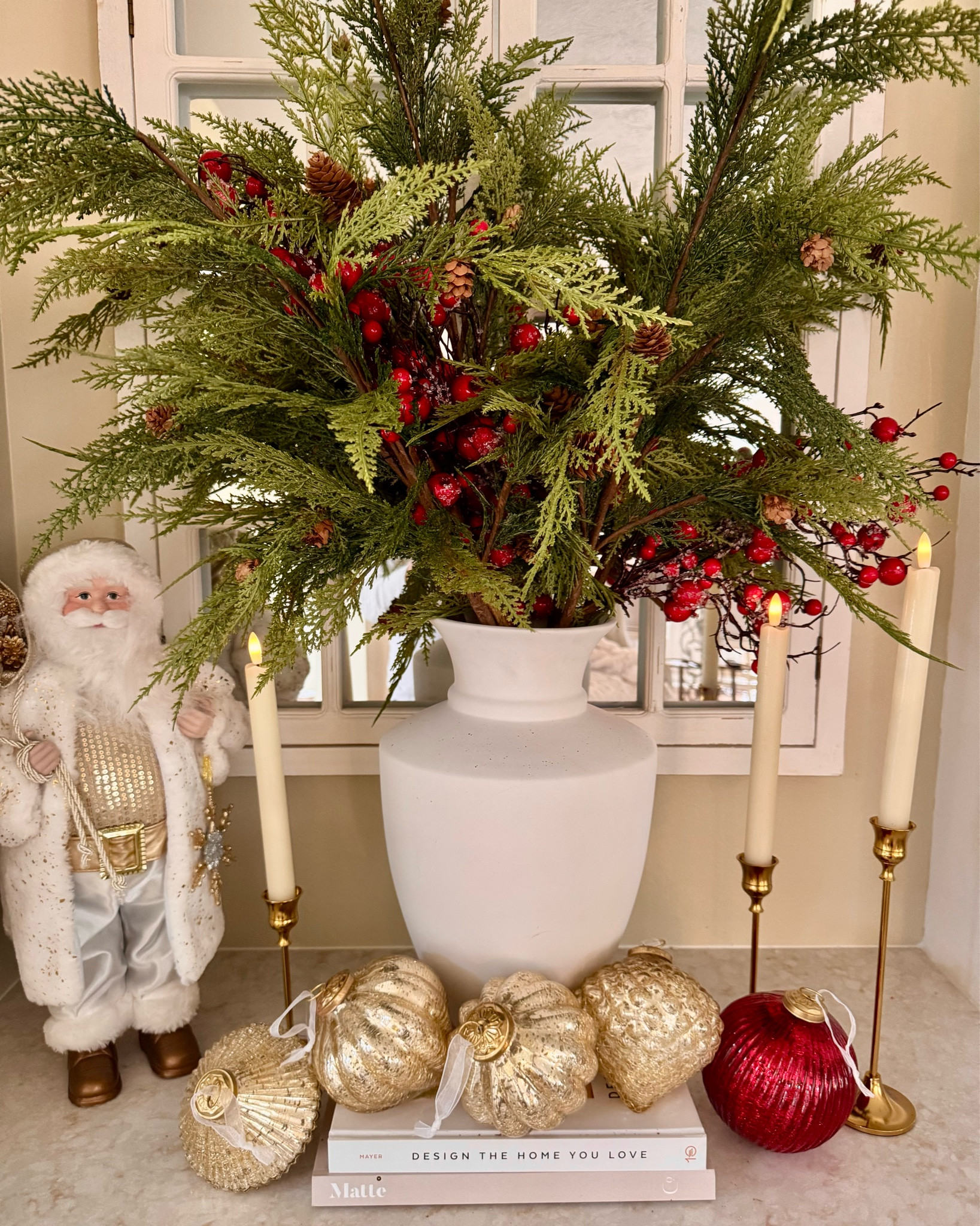 Elegant Christmas vignette featuring a lush greenery arrangement with red berries and pinecones, complemented by a white vase, gold-accented ornaments, brass candleholders, and a charming Santa figurine, creating a cozy and festive holiday display.

#LTKHoliday #LTKSeasonal #LTKHome