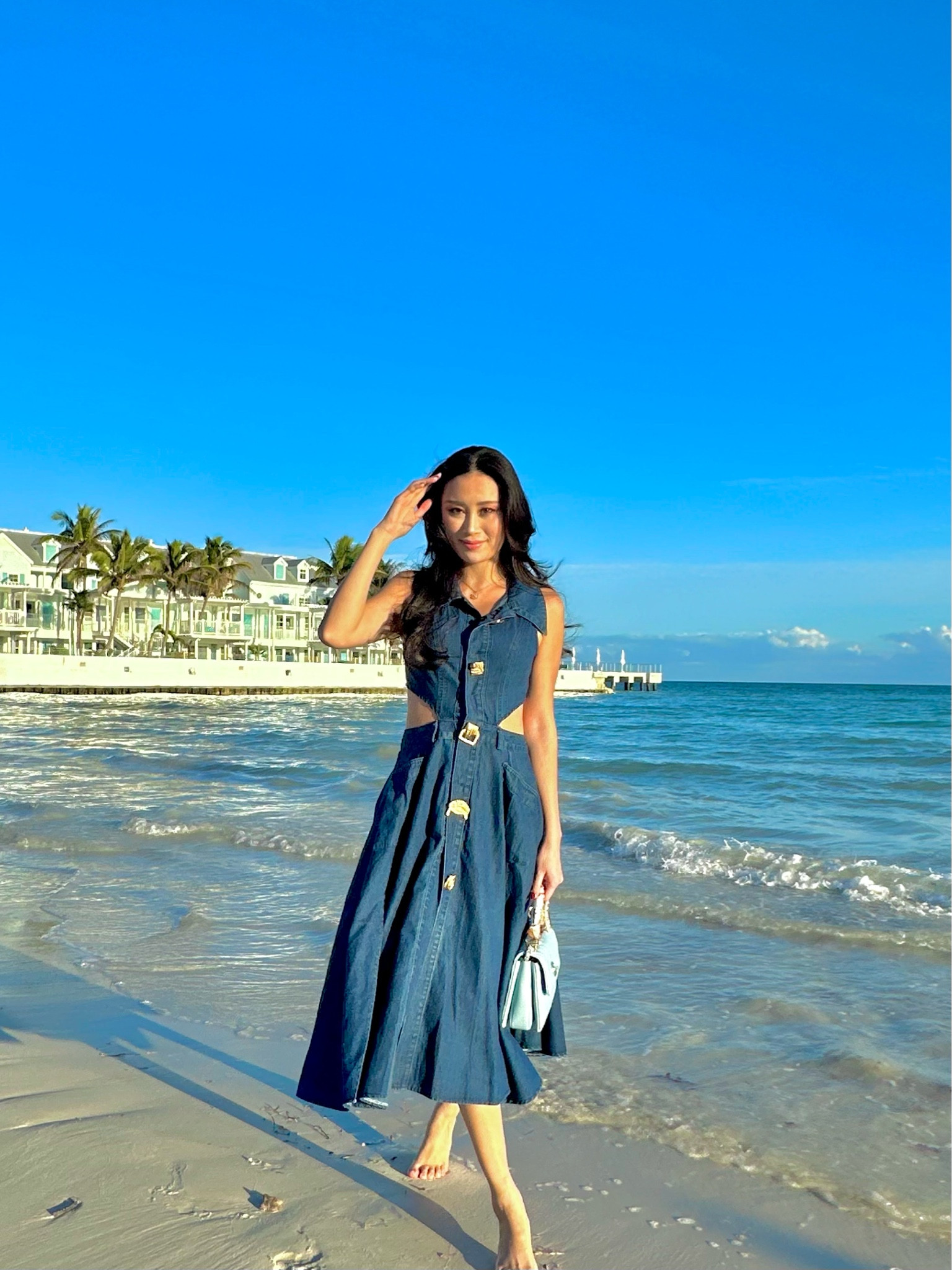 a denim midi dress with gold statement buttons, a fitted collared bodice, side waist cutouts, and a full a-line skirt with patch pockets — styled with a baby blue top-handle bag for a barefoot beach walk on a tropical coastal getaway. the elevated denim and gold hardware make this the perfect young old money resort dress. perfect for key west, the bahamas, palm beach, harbour island, and east coast island vacations. shop this look linked above.