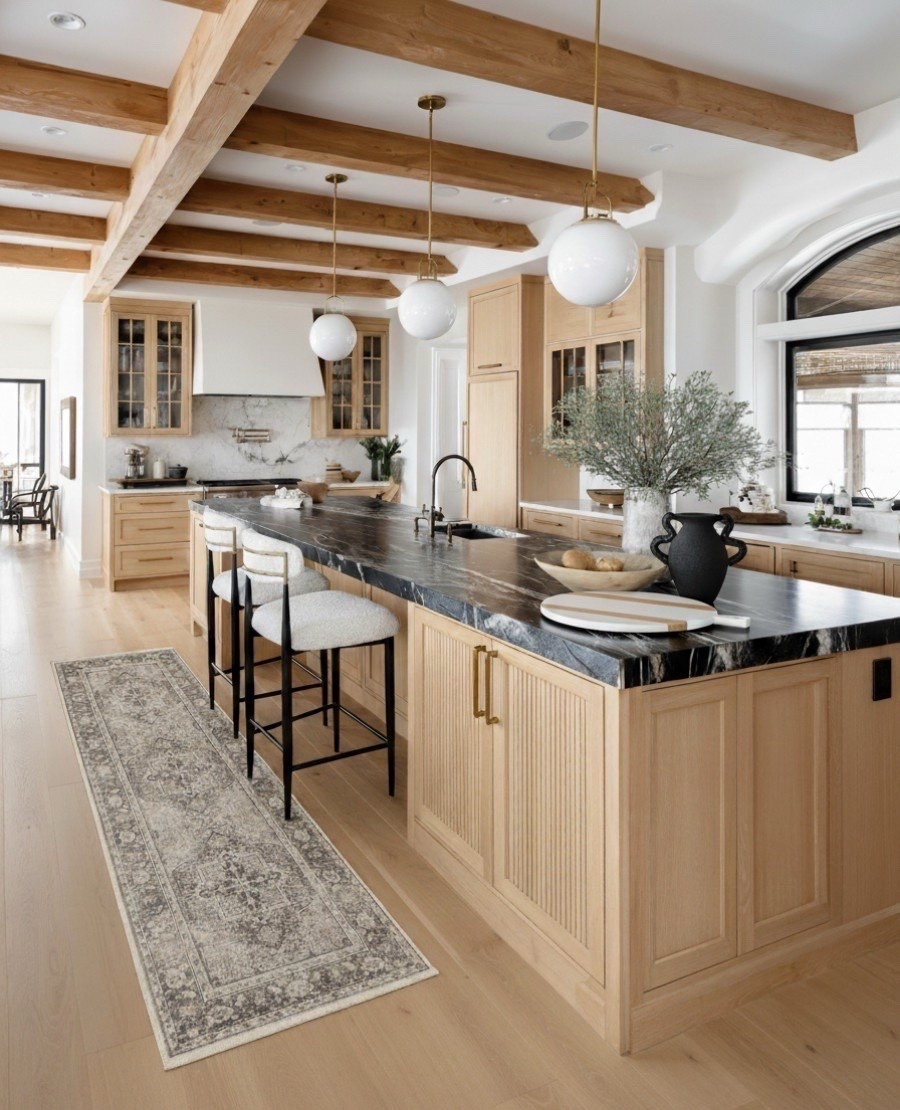 A bright and welcoming kitchen that mixes natural warmth with elevated modern details. The space is anchored by a light oak island with ribbed paneling, topped with a dramatic black marble countertop featuring soft white veining. The contrast creates a refined focal point that feels both organic and luxurious. Overhead, white globe pendants with brass accents bring a soft glow and a touch of classic charm.

Along the perimeter, light oak cabinetry keeps the palette airy while glass front uppers add a curated feel. The marble backsplash in creamy white and soft gray ties in beautifully with the counters. A pair of upholstered counter stools in ivory fabric with brass trim and matte black legs introduces a clean tailored look.

The vintage style runner in tones of taupe, charcoal, and ivory adds warmth and pattern underfoot. Layered decor such as the black ceramic vase, textured stone vessel, olive branches, and neutral serving pieces soften the hard surfaces and bring in earthy character. Exposed natural wood ceiling beams complete the space with rustic charm that feels perfectly balanced.

✨ Shop the look:
Light oak kitchen island | Black marble countertop | White globe brass pendants | Light oak cabinetry | Glass front upper cabinets | Marble backsplash | Ivory upholstered counter stools with brass detail | Black ceramic vase | Stone textured vase | Neutral serving bowls | Vintage taupe and ivory runner | Natural wood ceiling beams 



#LTKHome #LTKSaleAlert