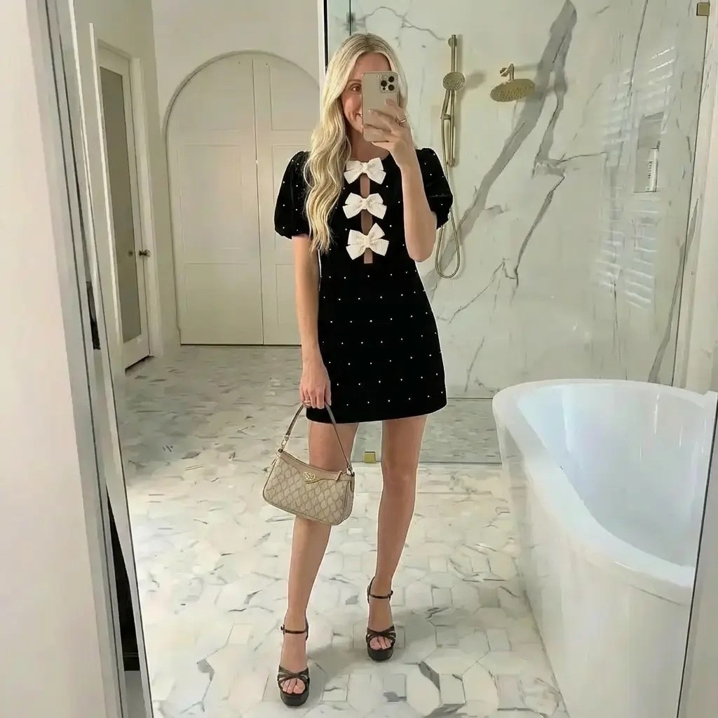 A refined mirror selfie in a bright, marble-clad luxury bathroom. A tall, elegant blonde woman wears a fitted black mini dress adorned with delicate white bow details down the front, short puff sleeves, and subtle sparkle accents. She pairs the look with black strappy heels and a small beige designer shoulder bag. Her hair is styled in loose, glossy waves, and the lighting is soft and natural, highlighting a clean, minimalist, high-end aesthetic. The scene feels feminine, sophisticated, and effortlessly chic — a modern quiet-luxury look with Parisian-inspired elegance.

⸻

HASHTAGS

#QuietLuxury
#FeminineChic
#BowDress
#LittleBlackDress
#ClassicWithATwist
#EffortlessElegance
#ParisianVibes
#LuxuryAesthetic
#ChicStyle
#MirrorOutfit
#MinimalistFashion
#ModernRomantic
#TimelessStyle
#BlackDressMoment
#ElegantDetails
#FashionInspo
#NeutralLuxury
#SoftGlam
#OldMoneyAesthetic
#OOTD

#LTKHoliday #LTKdayinmylife #LTKGiftGuide