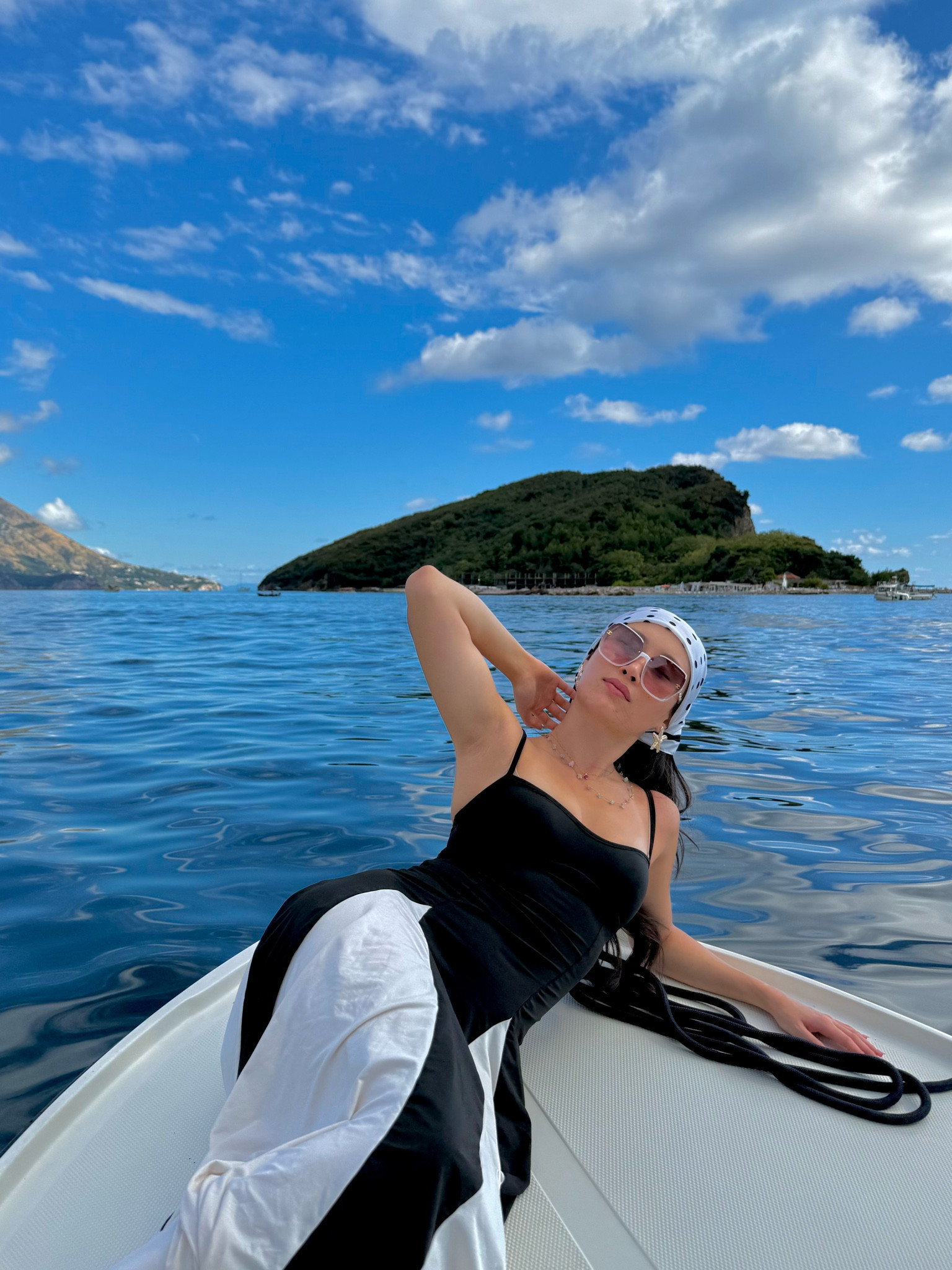 a black and white halter maxi dress with a fitted bodice, fluid bias-cut silhouette, and contrast color blocking, styled with a white polka dot silk headscarf, oversized rose-tinted square sunglasses, and layered delicate necklaces for a yacht day on the budva coast with sveti nikola island in the distance. the graphic black and white contrast and elegant maxi silhouette make this the ultimate mediterranean boat day dress. perfect for yacht days, mediterranean cruises, balkan coast vacations, and montenegro boat trips. shop this look linked above.