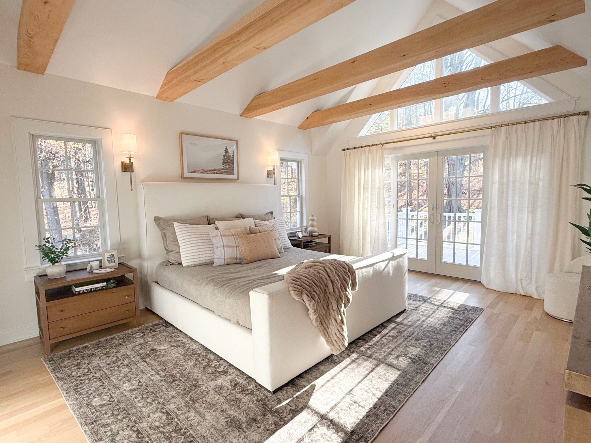 A stunning view of the Mount Kisco primary bedroom reveals sliding glass doors that open to a private balcony, filling the space with natural light and a connection to the outdoors. The vaulted ceiling, accented by exposed wooden beams, adds architectural interest and a sense of spaciousness. The room’s soft textures and neutral palette create a cozy yet airy retreat. 
