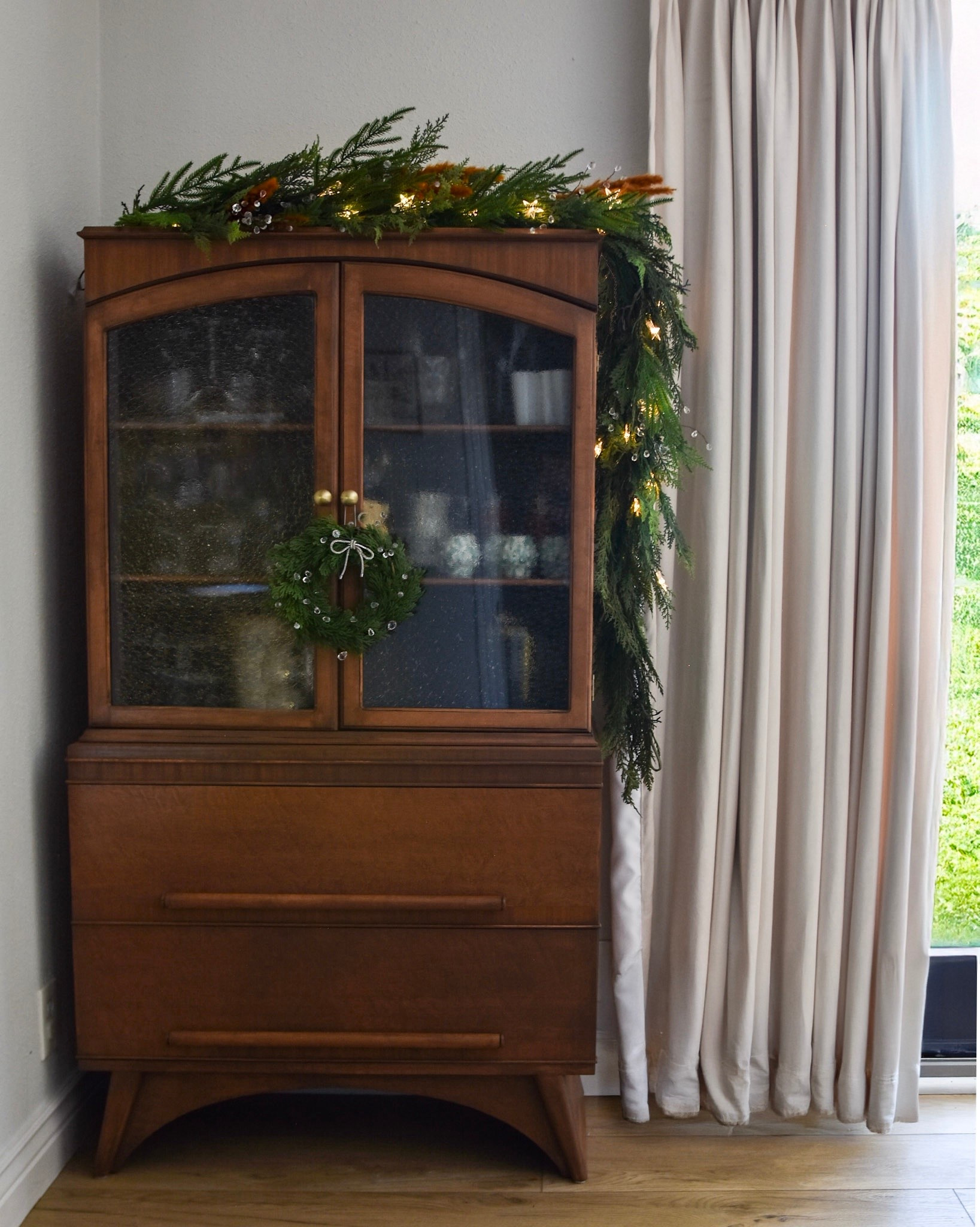 Vintage hutch revival: complete. ✨
What started as a tired little cabinet is now a warm mid-century moment in the corner of our living room—fresh glass doors, clean lines, and dressed up with garland + twinkle lights for the Christmas season. The rich wood, textured glass, and simple brass knobs do most of the talking, the greenery is just the finishing touch! 

#hutchmakeover #hutchrevival #midcenturymodernhome #vintagecabinet #furnitureflip #diyhomedecor #christmasdecor #holidayhutch

#LTKHome #LTKHoliday