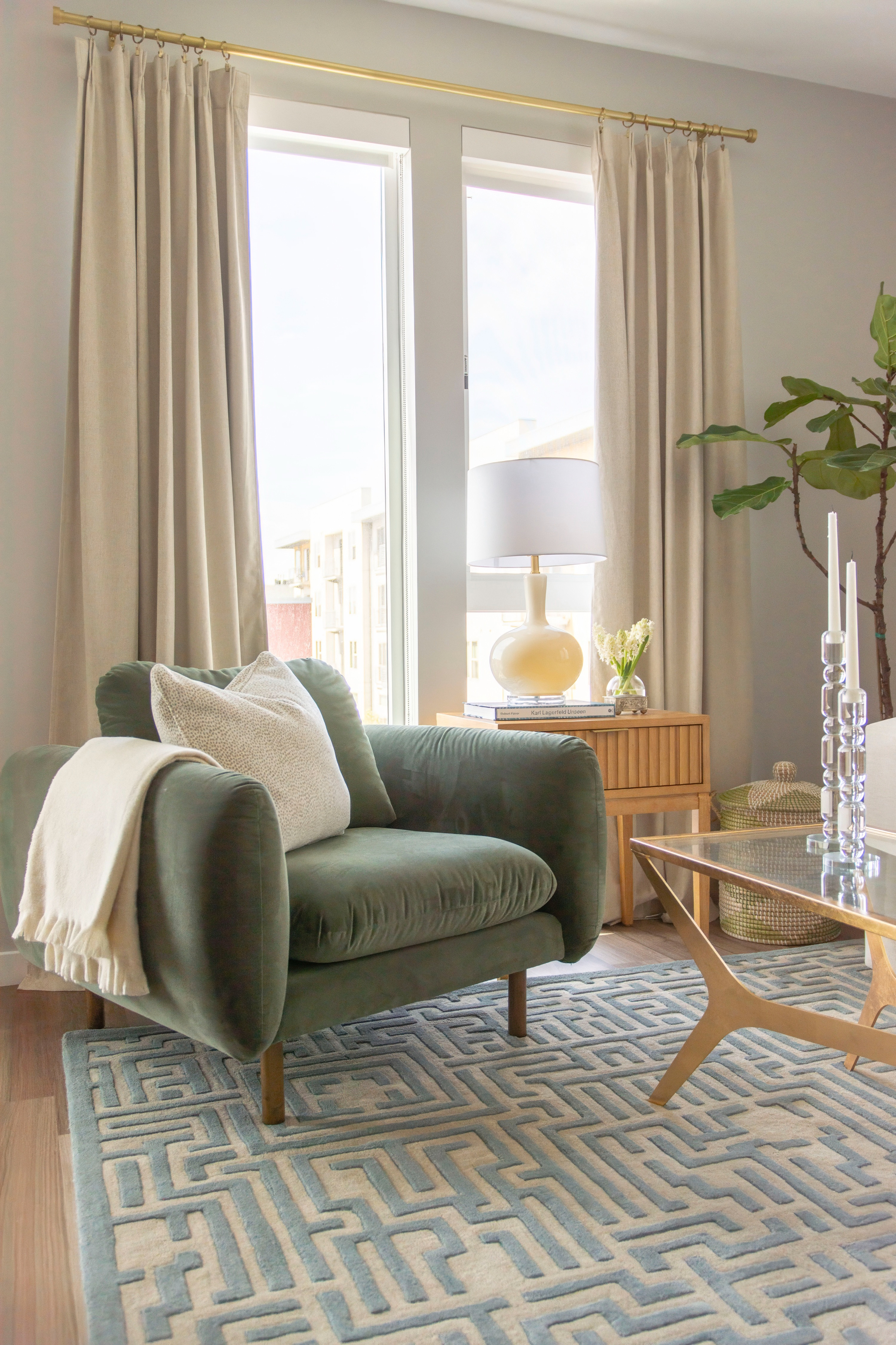 Cozy modern living room design with a chic mix of textures and neutrals. Featuring a plush sage green velvet armchair styled with a soft ivory throw and textured pillow, layered over a geometric blue and cream area rug. Natural light filters through neutral linen curtains on a brass curtain rod, complementing the mid-century inspired wood side table with a ceramic table lamp and fresh florals. A glass coffee table with gold legs adds a touch of modern glam, while a tall indoor plant brings in greenery. Perfect inspiration for apartment living, cozy corners, and modern home decor styling. 

 