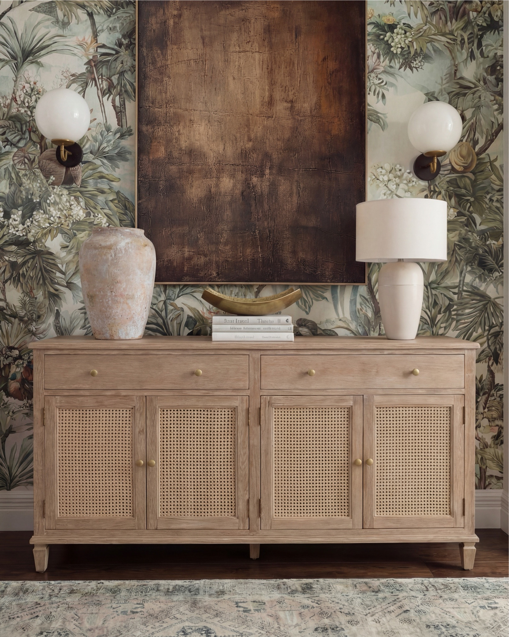 There’s something about a moody, nature-inspired foyer that just sets the tone for the whole home, and this one does it so effortlessly. The mix of warm wood, soft texture, and bold art makes the space feel collected and elevated from the moment you walk in.

The natural-oak console brings the perfect balance of clean lines and subtle detail with its woven cane doors. I styled it with an oversized stone vase, a tall ceramic lamp, and a sculptural gold bowl layered on books — just enough to feel intentional without being cluttered.

The vintage-inspired runner adds muted pattern and softness underfoot, grounding all the warm wood tones beautifully. And that wallpaper — the deep greens, palms, and floral textures make the entire wall feel like a piece of art on its own.

The oversized copper-toned artwork brings so much richness to the scene, pulling in the darker notes from the sconces and the natural shadows in the wallpaper. It’s the kind of statement piece that instantly elevates a foyer.

✨ Shop the Look
Natural-oak cane console | Oversized stone vase | Sculptural gold bowl | Ceramic table lamp | Botanical wallpaper | Vintage-style runner | Copper-toned oversized artwork | Globe wall sconces

#LTKhome #LTKsale #LTKstyletip