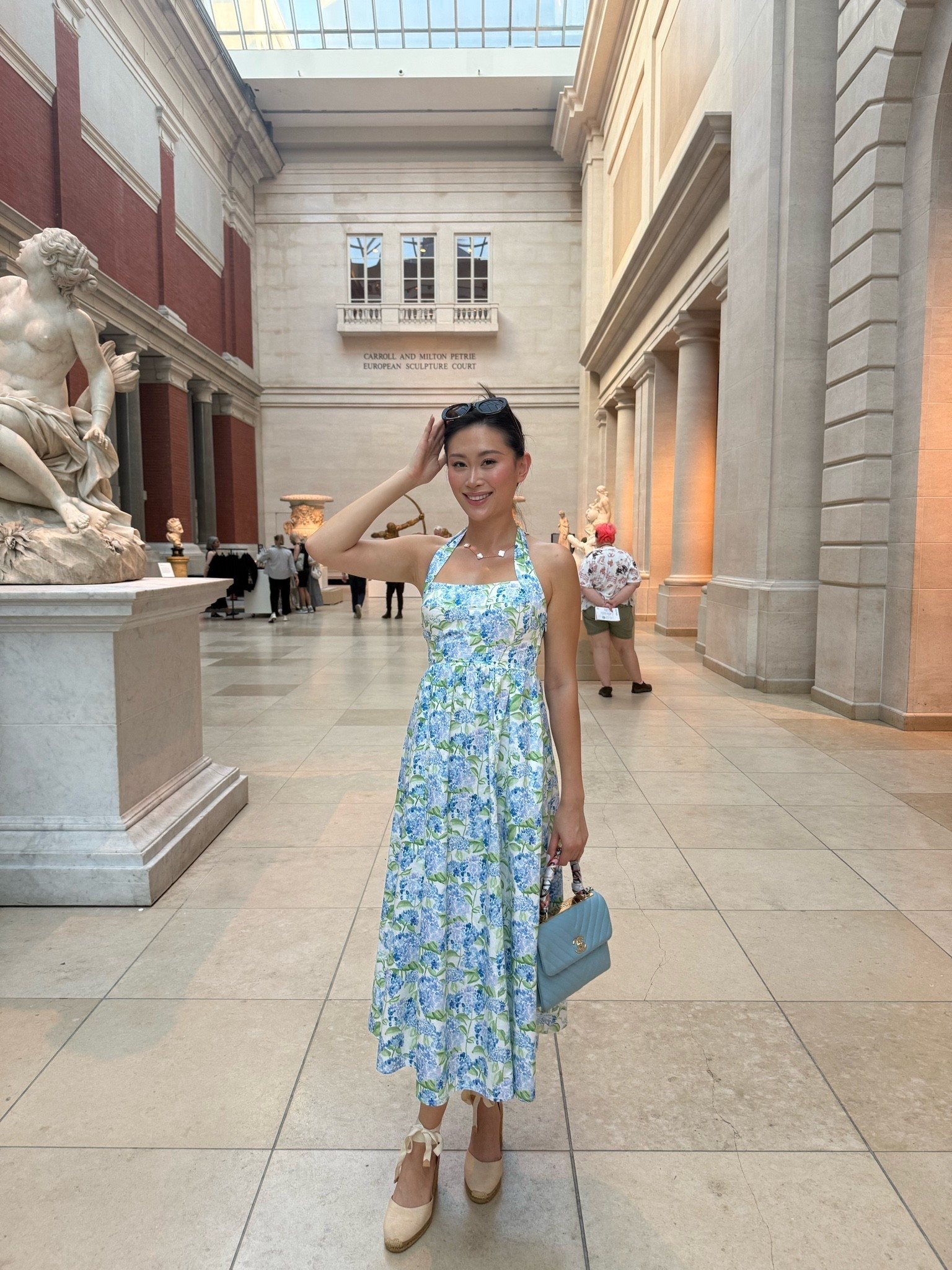 a Hill House blue hydrangea floral print halter midi dress with a fitted bodice and full skirt, styled with cream ribbon-tie espadrille wedges, a powder blue quilted top-handle bag, and oval sunglasses. the hydrangea print and halter silhouette make this an instant warm-weather favorite that reads polished whether you’re at a museum or a garden lunch. perfect for city day dates, museum visits, garden parties, and summer weddings. shop this look linked above.