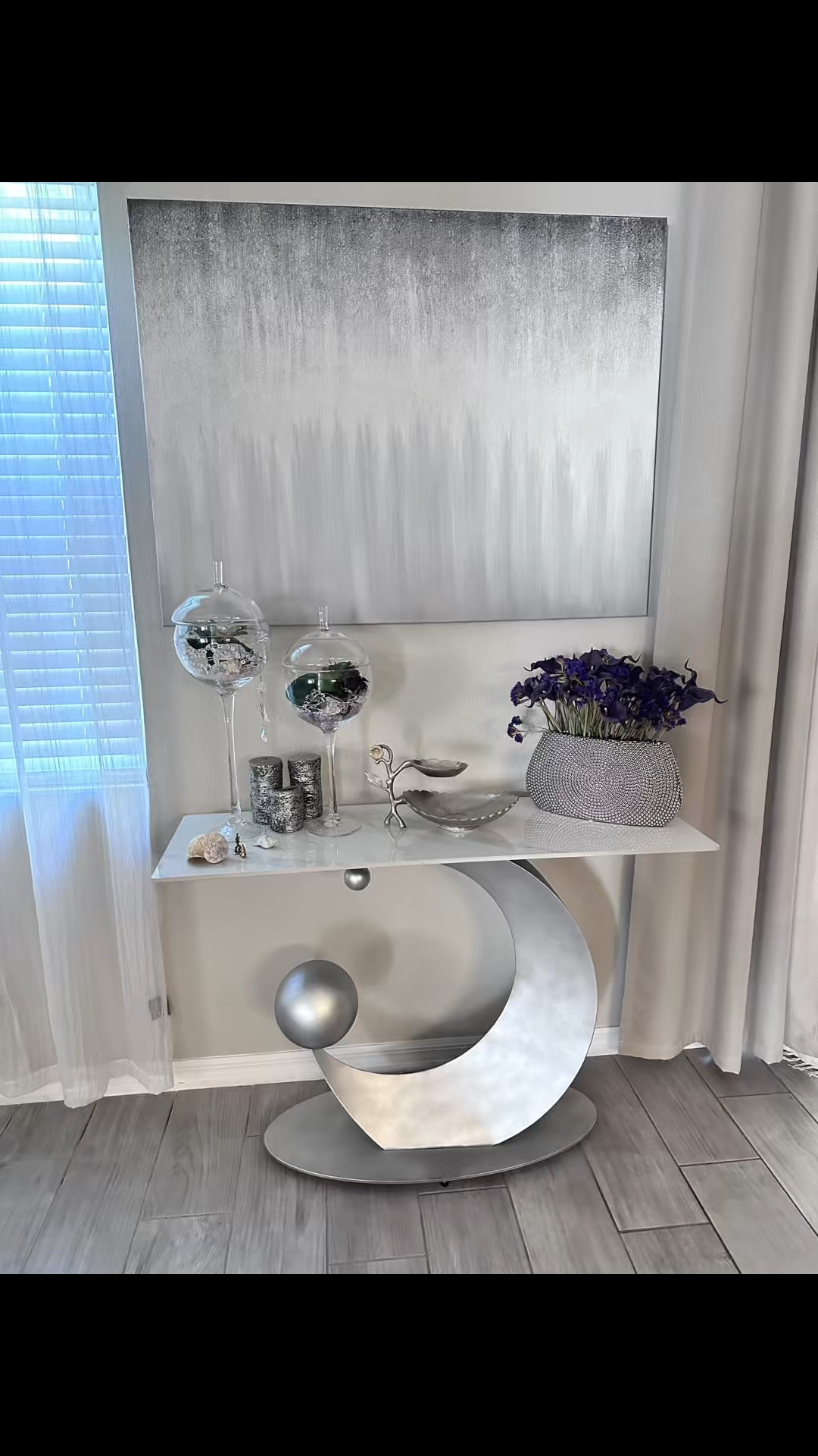 ✨ Modern Elegance for Your Home ✨

Creating a cozy yet sophisticated space is all about mixing textures and unique pieces, and this setup captures that perfectly!

🌟 Featured Pieces:
	•	Statement Console Table: This artistic silver crescent design with metallic accents is a true showstopper. (Just for info I painted this console in silver to match my interior designs in my house original color in the shopping list)
	•	Glass Terrariums: Add a touch of greenery and natural beauty with these elegant hanging glass planters.
	•	Iridescent Accessories: The shimmering candle holders and metallic bowls bring a touch of glamour.
	•	Floral Arrangement: Deep purple blooms in a textured vase add color and contrast to the neutral tones.
	•	Abstract Artwork: The gradient gray canvas ties the entire look together with its minimalist charm.

💡 Styling Tip: Balance bold statement pieces with soft curtains and natural lighting to keep the space inviting and bright.

🛍️ Shop similar home decor pieces through my LTK profile. Let’s make your home a masterpiece! #HomeDecorGoals #ModernLiving #LTKHome