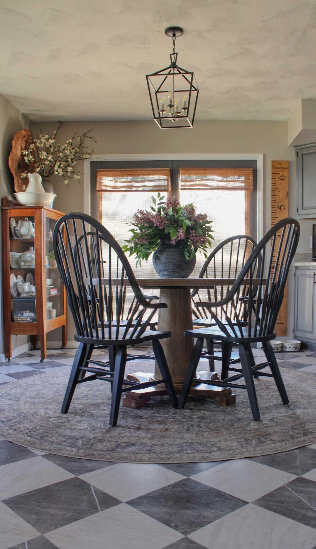 A moody vintage-modern kitchen featuring a round solid wood pedestal table, Windsor solid wood dining chairs, and an accent cabinet full of antique serve ware. Spring florals serve as a colorful centerpiece to this cozy, neutral aesthetic. A round machine washable rug grounds the space and the double doors add depth and dimension to this farmhouse inspired dining room.

#LTKHome