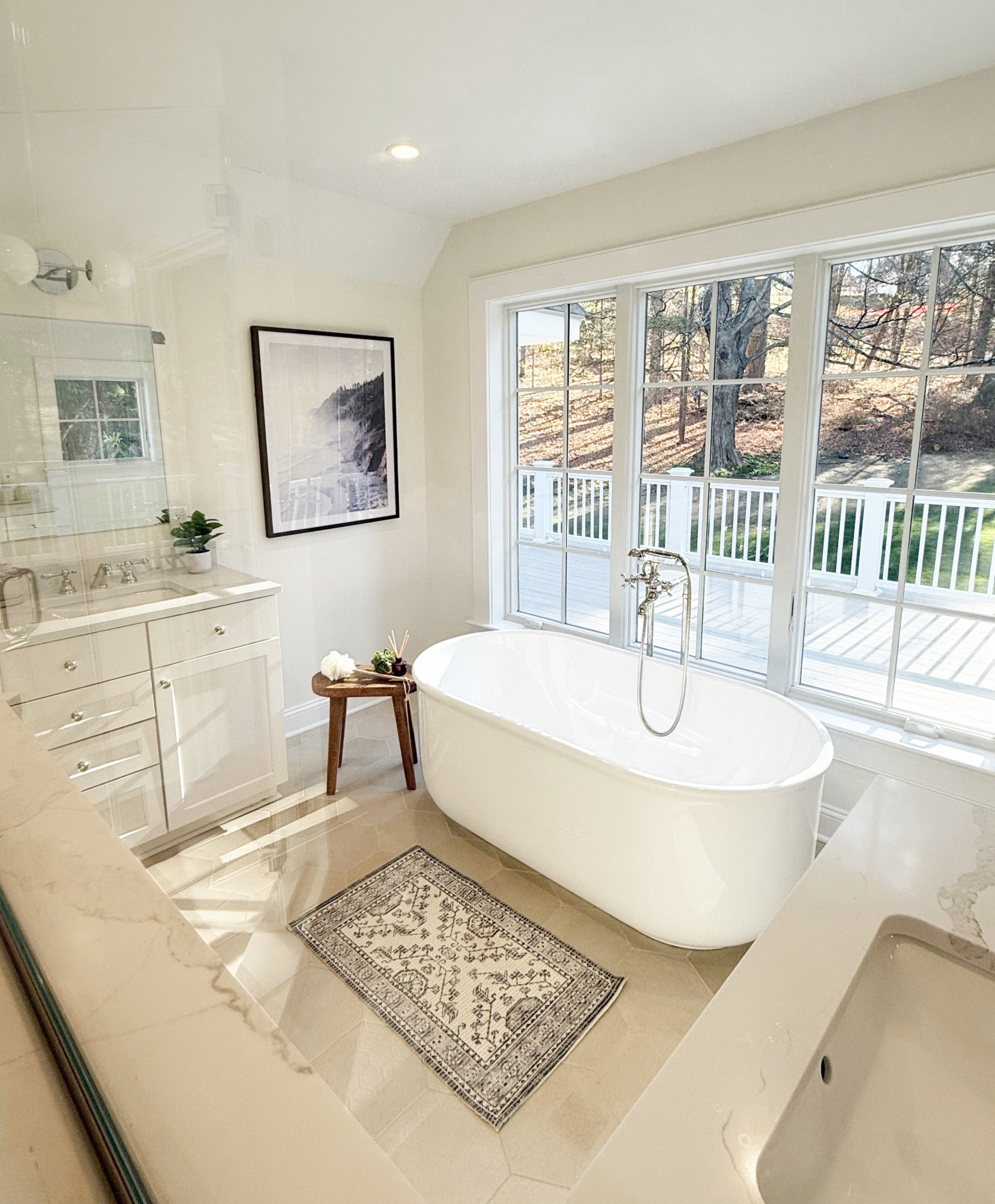 Another tranquil view of the Mount Kisco primary bathroom showcases the freestanding tub nestled beneath a large window, inviting natural light to pour in. A simple wooden stool adds a touch of warmth and functionality, while one of the matching white vanities offers a glimpse of elegant marble countertops and sleek hardware. The serene palette and thoughtful layout make this space feel like a luxurious at-home spa.

