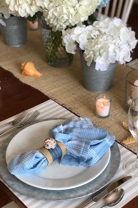 Summer table setting with galvanized charger plates, white dishes, blue napkins and white hydrangeas.  Coastal style with shells, candles and a burlap table runner. 

#LTKHome #LTKSeasonal #LTKStyleTip