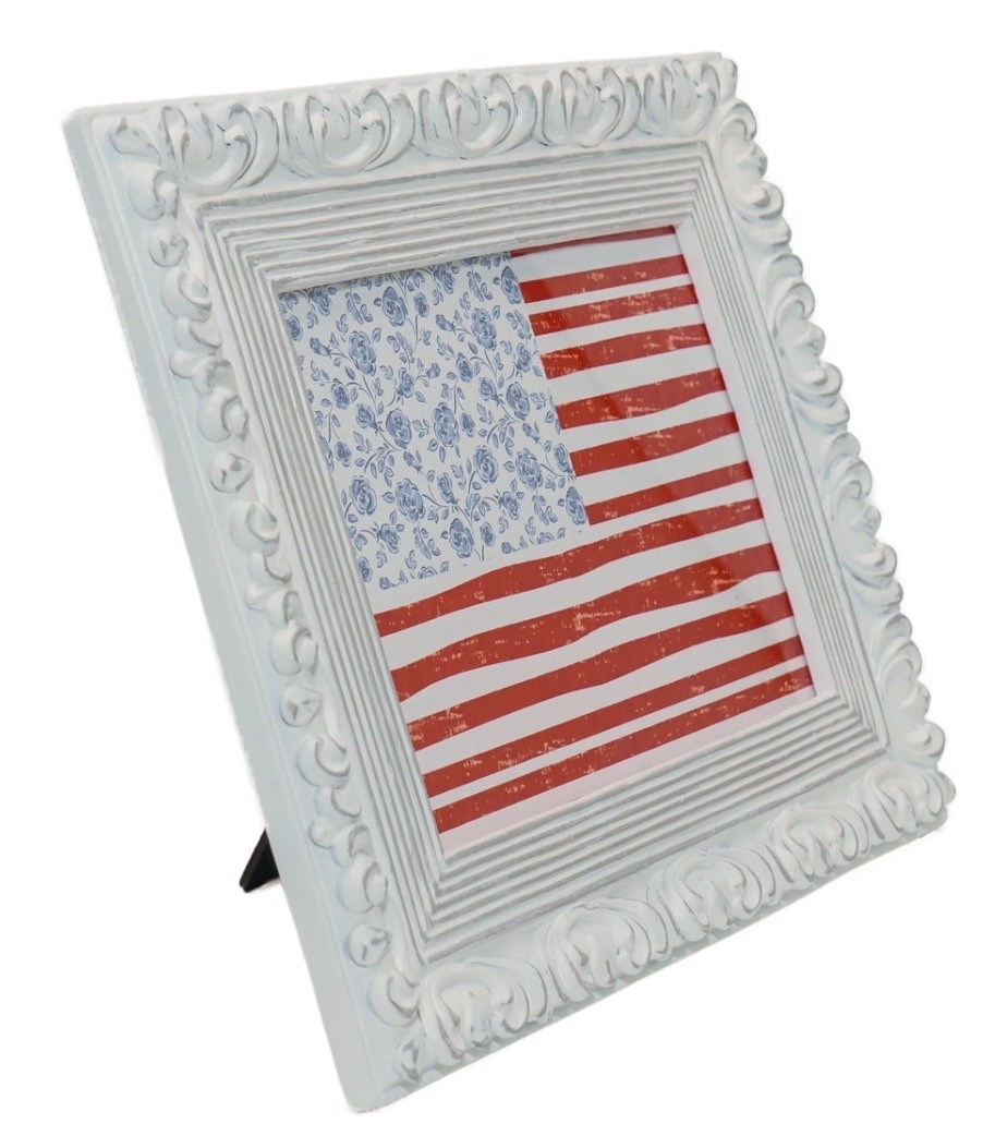 Add a touch of classic, decorative style to your home with the My Texas House Floral Flag Hanging Sign. This beautiful piece features a stylized American flag design housed within an ornate, white-washed frame with intricate scrollwork details. The artwork itself has a charming, rustic feel, with weathered red and white horizontal stripes. In a unique, refined twist, the top left corner features a white field adorned with a delicate blue floral pattern. The art hangs from a wide, light blue ribbon, tied in a lovely bow at the top, completing the soft and inviting look. This piece is a perfect way to bring an effortlessly stylish and patriotic touch to any room.