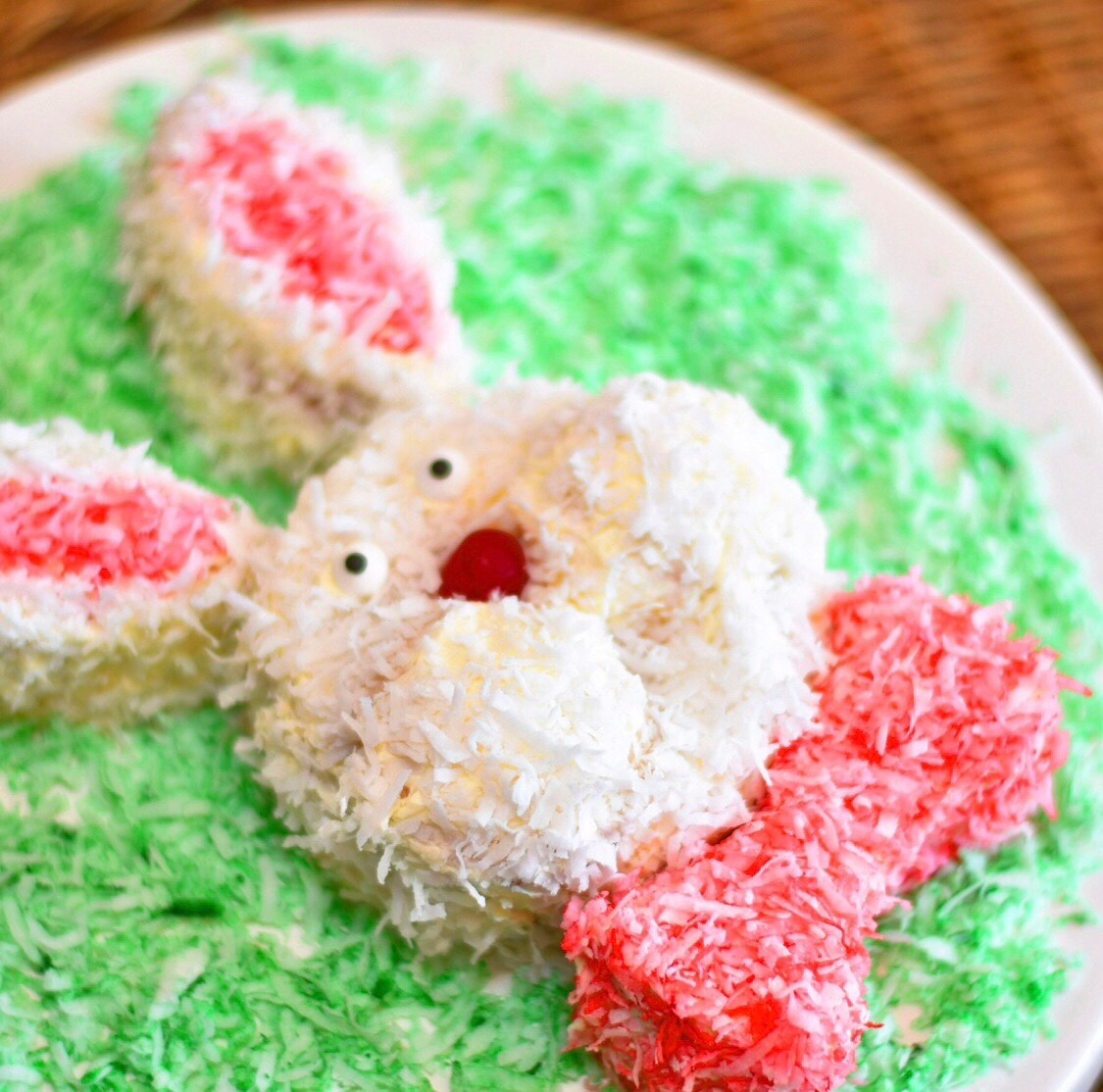 Following the directions on the box of the cake mix you are using, bake 2 round cakes and 2 cupcakes. I find using spring form pans for the round cakes work best. I used 3 inch spring form pans instead of 9 because I wanted a smaller cake and I made Walker cupcakes with the remaining batter.
Bake as directed and cool.
Once cooled, cut 2 convex shaped ears from each side of one round cake, creating 3 pieces- two ears and a bow tie.
Next, cut the tops off the cupcakes. You’ll only need the tops so feel free to eat the bottom halves- quality control is key!
On your cake stand or serving tray, frost the whole/uncut round cake with your favorite white frosting. This will be the bunny’s “head”.
Place the cupcake tops onto the round cake side by side.  These are the “cheeks” of your bunny.  Frost the tops of the cupcakes.
Next place the “ears” and “bow tie” on your serving try and frost.
I like using food coloring to create colored coconut for the inside of the ears and the box tie. To do this place coconut in a ziplock bag with a few drops of desired color food coloring.  Seal and move the coconut around in the bag until the flakes are coated evenly.
Sprinkle white coconut on your bunny and decorate with colored coconut and candies for “eyes” and “noes”.

