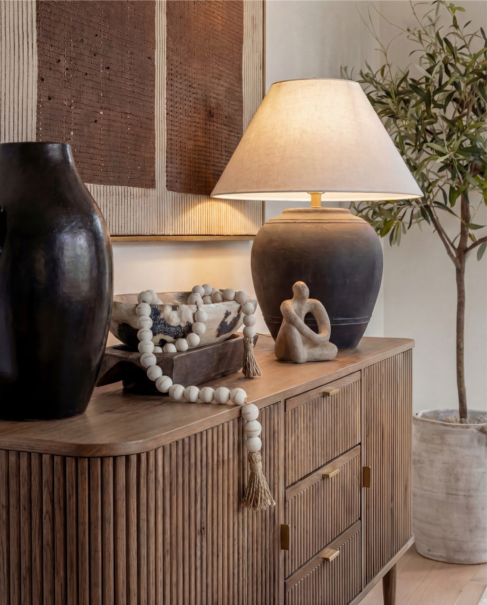 Shop the Look:
• Solid wood pedestal dining table (warm oak tone)
• Light wood dining chairs with upholstered seats
• Neutral vintage-style area rug (soft brown pattern)
• Brass chandelier with tapered fabric shades
• Exposed wood ceiling beams
• Large wood and glass display cabinet
• Fluted wood sideboard cabinet with brass hardware
• Black ceramic table lamp with linen shade
• Framed neutral textile wall art
• Matte black decorative vase
• Wood bead garland with tassels
• Decorative wood tray with ceramic bowl
• Small abstract sculpture decor
• Olive tree in stone planter
• White scalloped ceramic centerpiece bowl
• Dark ceramic pitcher vase (table decor)
• Glass hurricane candle holder
• Sheer neutral curtains



#LTKHome #LTKSaleAlert