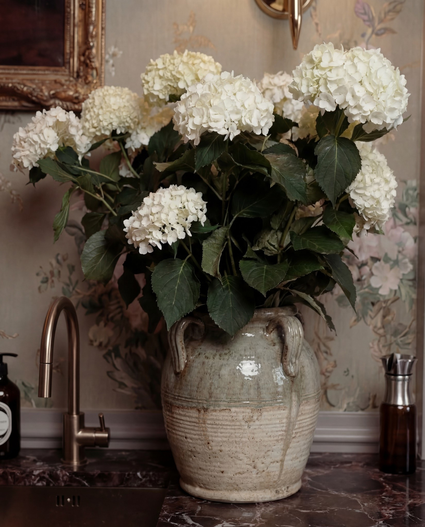 Shop the Look:
• Custom bar cabinetry (charcoal or deep green, reeded fronts)
• Brass cabinet pulls and knobs
• Dark marble or stone countertop (brown, burgundy, or black veining)
• Floral wallpaper (muted green, taupe, blush tones)
• Brass wall sconces with pleated fabric shades
• Framed vintage artwork (ornate gold frame)
• Smeg Espresso machine (cream or off-white)
• Ceramic mug or glass cup
• Wooden tray for bar essentials
• Glass bottles and barware
• Stoneware vase (aged or textured finish)
• White hydrangea floral arrangement
• Built-in sink with brass faucet
• Neutral vintage-style runner rug

#LTKSaleAlert #LTKFindsUnder100 #LTKHome