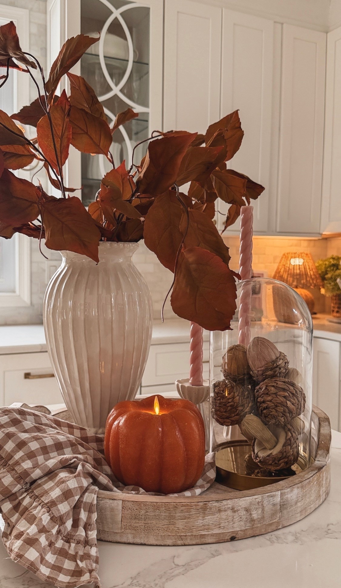 Fall kitchen island centerpiece with rust-colored branches in a white vase, orange and brown pumpkins, pink taper candles and a brown gingham cloth arranged on a round wood tray.
