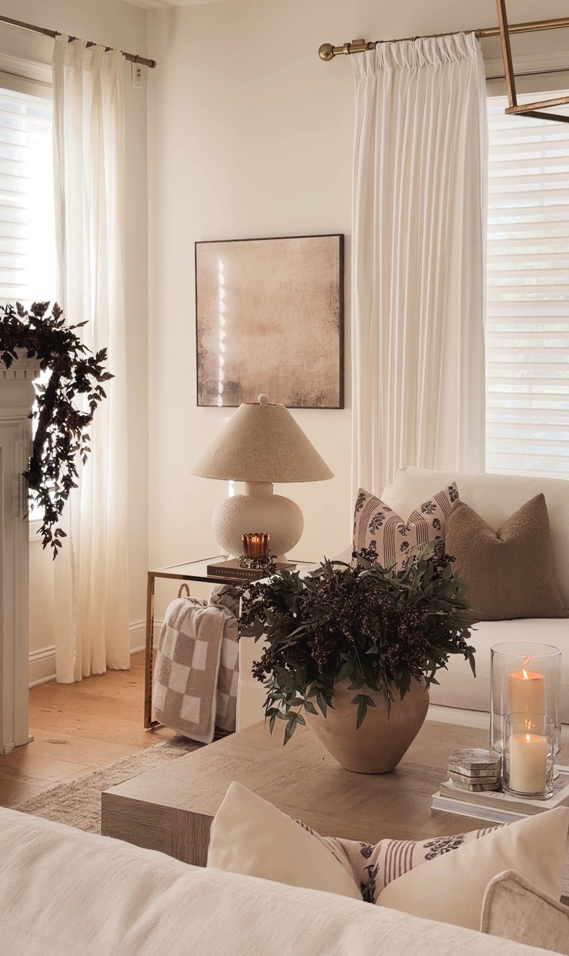 Neutral wood coffee table styled for fall with stacked design books, glowing glass hurricane candles, stone coasters, a white scalloped bowl filled with gold decorative pears and a large vase of dark greenery.
