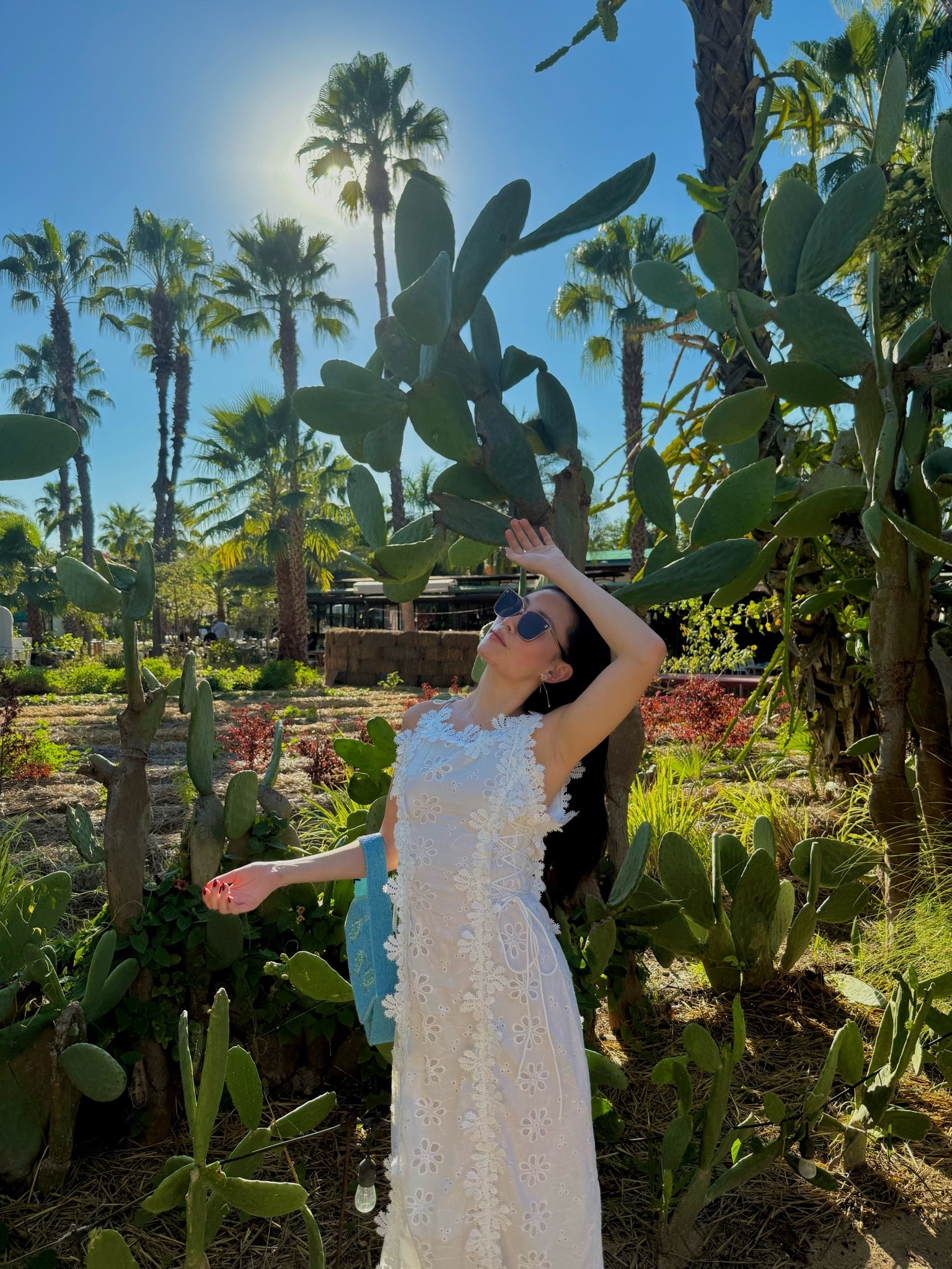 a white eyelet lace maxi dress with floral appliqué trim and a sleeveless silhouette, styled with a blue beaded top-handle bag and slim sunglasses. the broderie anglaise detailing and column shape make this the kind of white dress that photographs beautifully and works for every warm-weather event on the calendar. perfect for resort vacations, garden parties, summer brunches, and destination travel days. shop this look linked above.