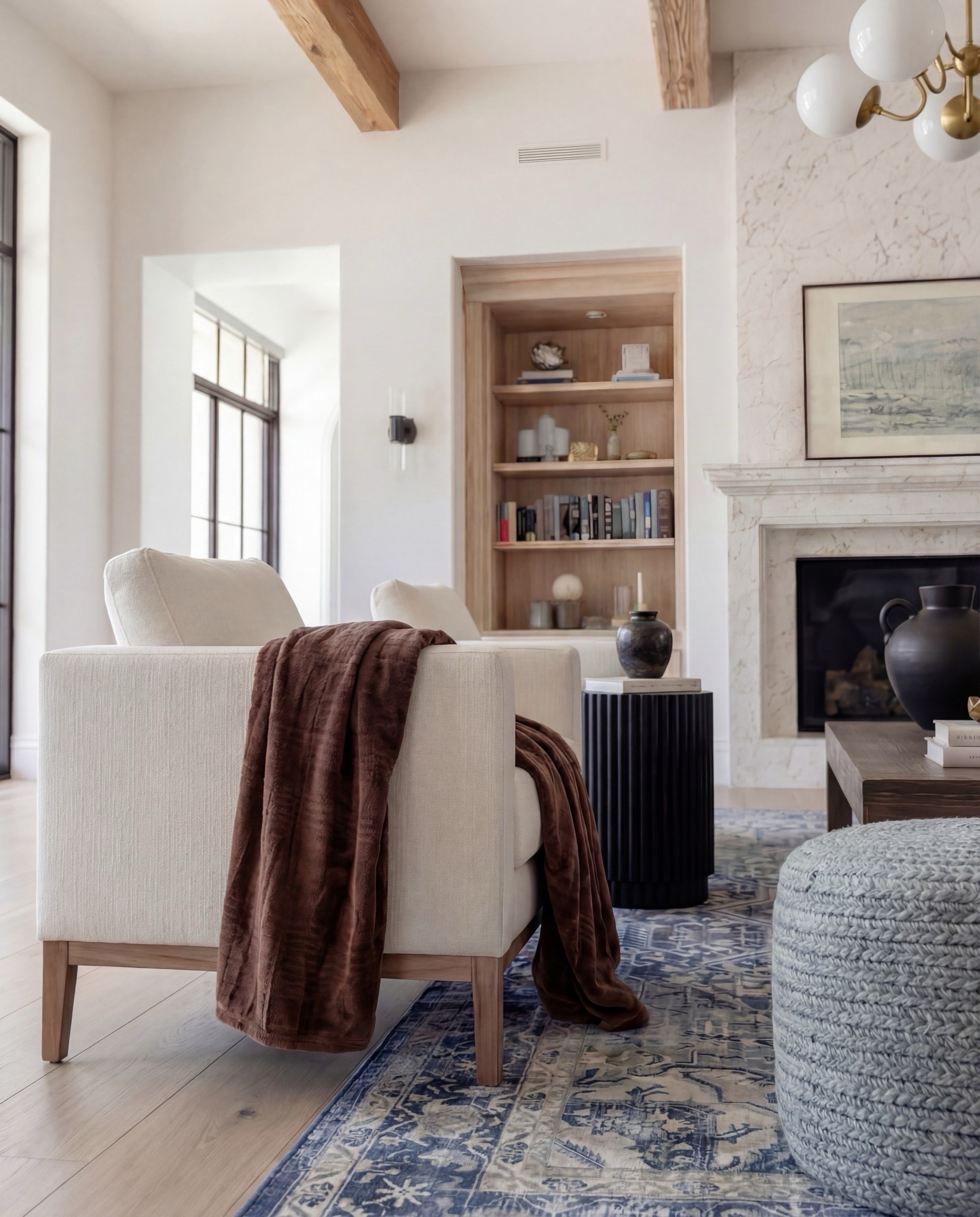 Shop the Look:
• Marble fireplace surround
• Cream armchairs + sofa
• Blue patterned lumbar + throw pillows
• Chunky knit ottomans
• Solid wood coffee table
• Black ceramic vase decor
• Distressed blue traditional rug
• Wood built-ins with glass fronts
• Warm wood ceiling beams
Brown throw blanket 
White decor books
Brass oval tray
Black decor bowl
Rounded globe chandelier 

#LTKHome #LTKSpringSale #LTKSaleAlert