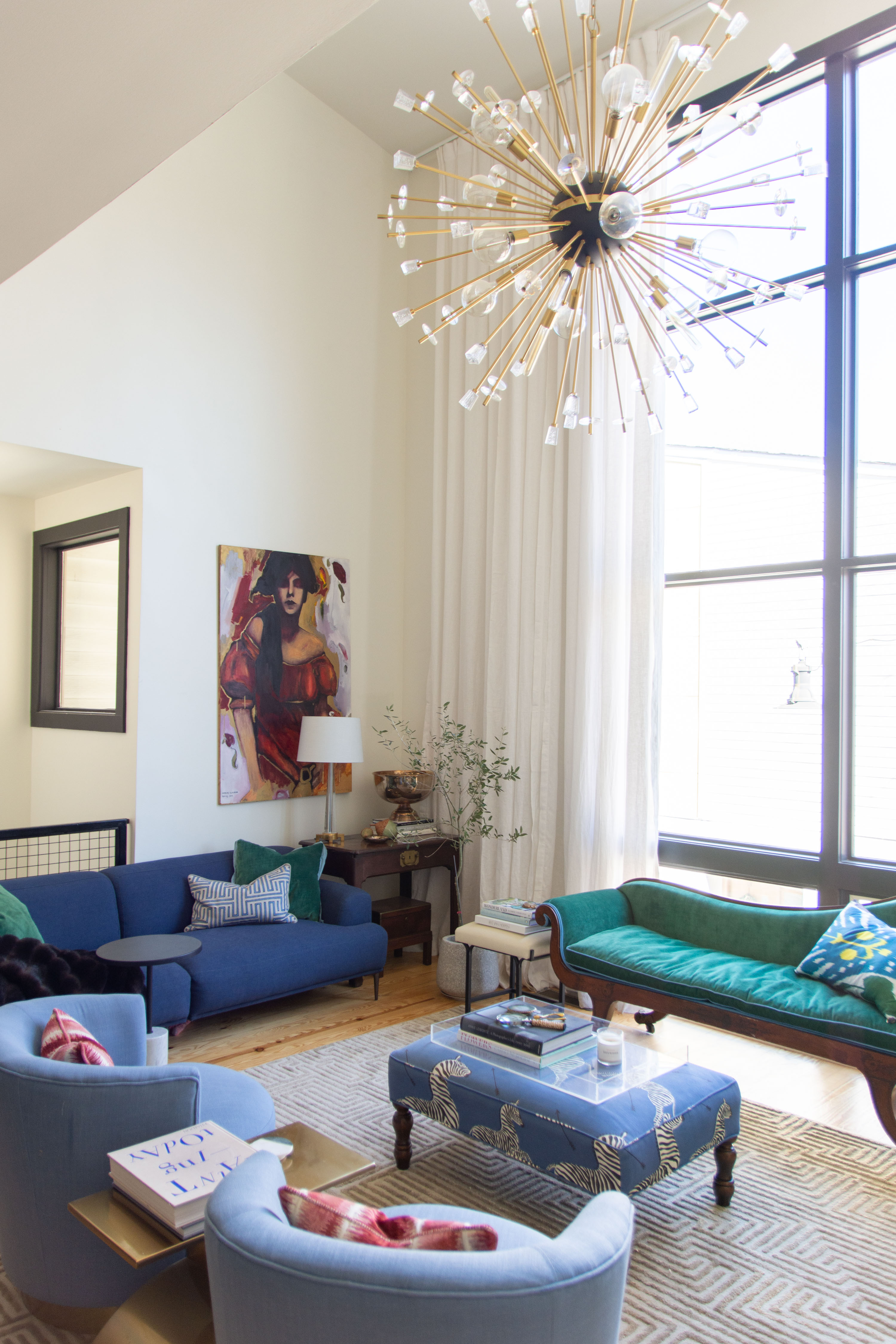 Modern eclectic living room design featuring a bold statement chandelier, mid-century inspired seating, and layered textures. This high-ceiling space combines jewel-tone velvet furniture with playful patterned accents for a chic yet inviting vibe. Blue armchairs pair with a navy sofa and a vintage green settee for a curated mix of styles. A zebra-print ottoman doubles as a coffee table, styled with books and trays for effortless decor. Large windows with floor-to-ceiling curtains flood the room with natural light, highlighting the gallery-style art and clean-lined rug. Perfect inspiration for colorful living room ideas, modern eclectic interiors, and statement lighting design. 

 