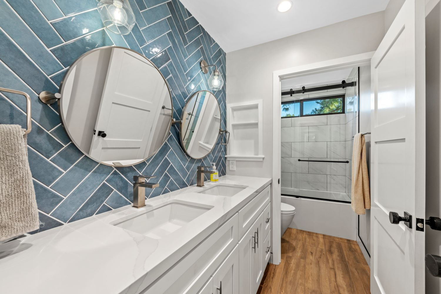 Dive into sophistication with our mesmerizing deep blue herringbone tile backsplash. Paired atop pristine white shaker cabinets and set against warm wood-like tile flooring, this design exudes contemporary elegance with a touch of coastal charm. Reflect your style with round pivot mirrors, adding depth and dimension to your space. 

#bathroomdesign #bathroombacksplash #backsplash #accenttile #accentwall #bluebathroom #bluetile  

 #LTKHome