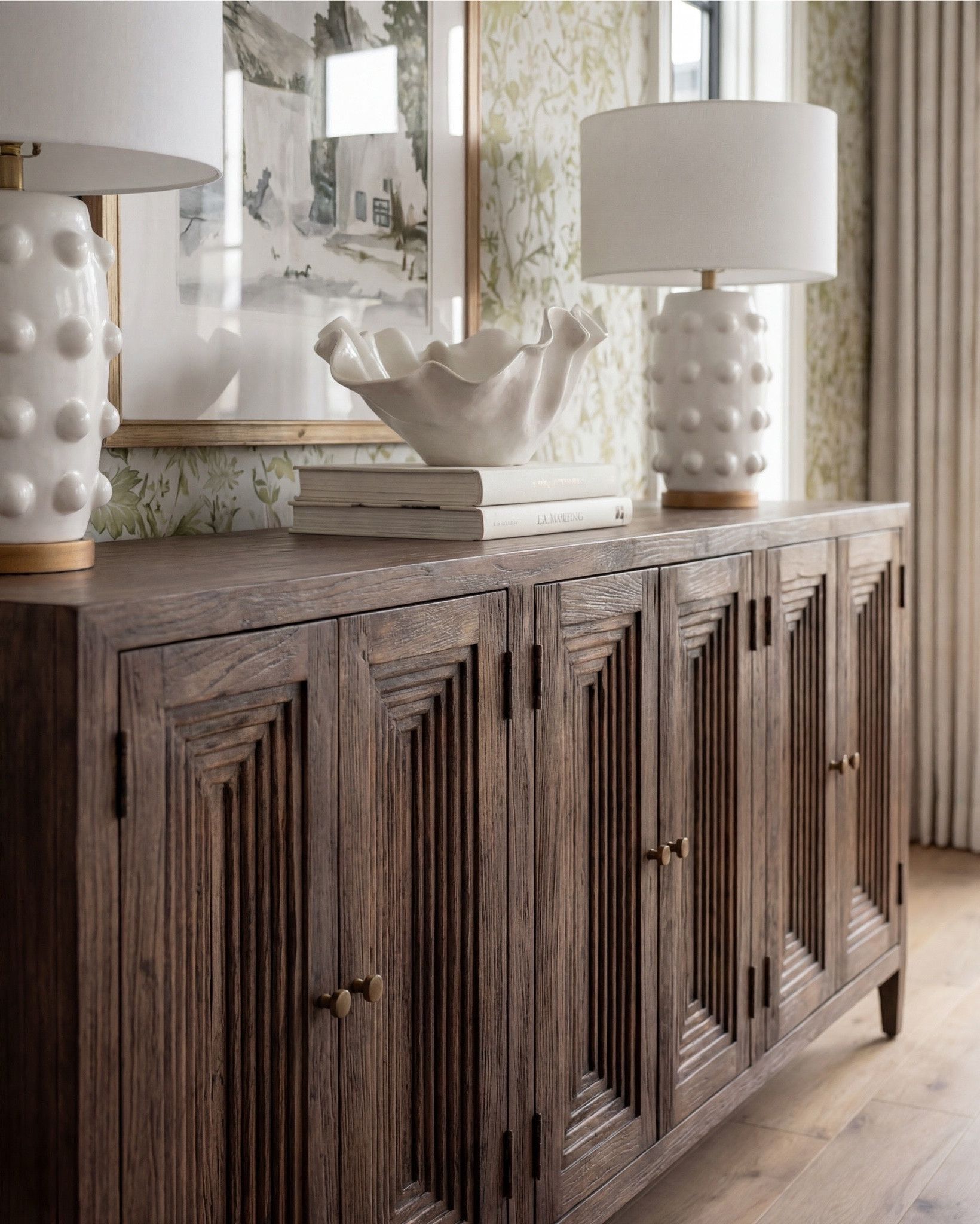 There’s something about a light filled, nature driven dining room that feels effortlessly welcoming the moment you step inside. This space blends soft pattern, warm wood, and organic lines in a way that feels both elevated and incredibly livable.

The pedestal dining table brings so much presence with its sculpted base and natural wood grain. I paired it with clean, curved dining chairs in a soft ivory upholstery to keep the look airy and balanced. On the sideboard, the deeper wood tone adds richness and grounds the entire room.

Styling is all about quiet impact here — a large ceramic vase filled with fresh greenery, a simple tray, and layered candles that keep the table looking intentional without taking away from the architecture around it. The twin lamps on the sideboard add symmetry and that perfect soft glow.

And that wallpaper — the muted green florals feel traditional in the best way, adding depth and texture without overwhelming the room. The dark, vintage inspired rug anchors the whole palette, pulling in those moody greens and creating contrast against the lighter woods.

✨ Shop the Look
Pedestal dining table
Curved wood dining chairs
Green floral wallpaper
Dark vintage inspired rug
Ceramic statement vase
Gold rim tray
Sideboard table lamps

#LTKhome #LTKsale #LTKstyletip