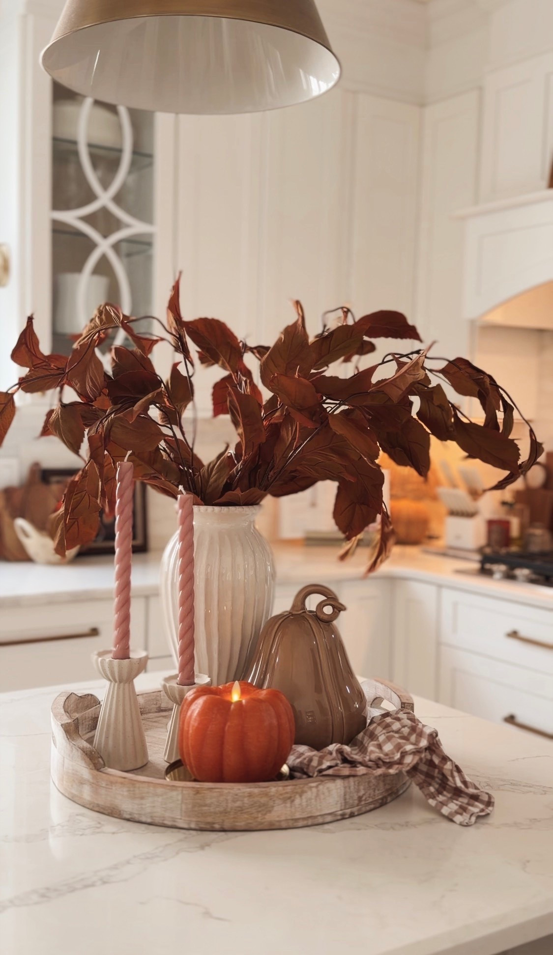 Close-up of an elevated fall kitchen centerpiece featuring a white ceramic vase with brown autumn branches, pumpkin candle, pink taper candle, woven acorns under a glass cloche and brown gingham fabric.