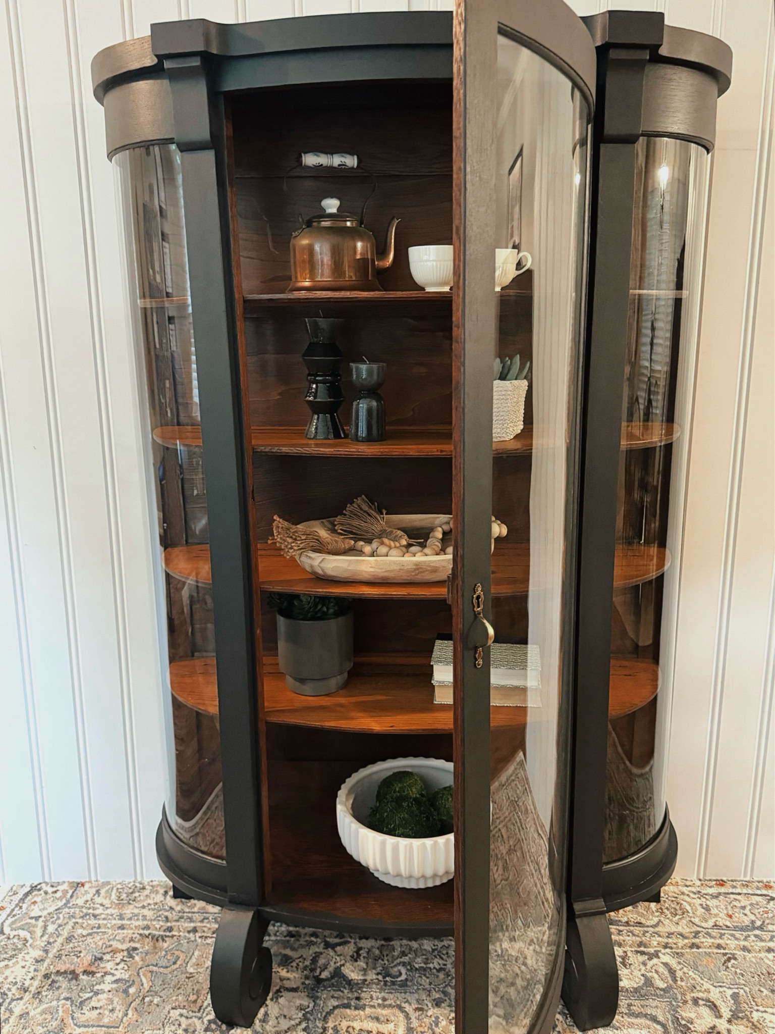This beautifully repainted display cabinet exemplifies how painting furniture can highlight its unique features and bring new life to a classic piece.

This antique display cabinet features a curved glass front with side panels, offering a panoramic view of its contents. The wooden frame has been newly painted in a dark finish, accentuating its elegant, rounded contours and sturdy build. The cabinet stands on short, curved legs, adding to its vintage appeal. Inside, the shelves provide ample space for showcasing various items, with the rich, dark wood interior creating a striking contrast against the lighter painted exterior. The cabinet door includes a decorative brass lock, enhancing its classic look.



#LTKHome