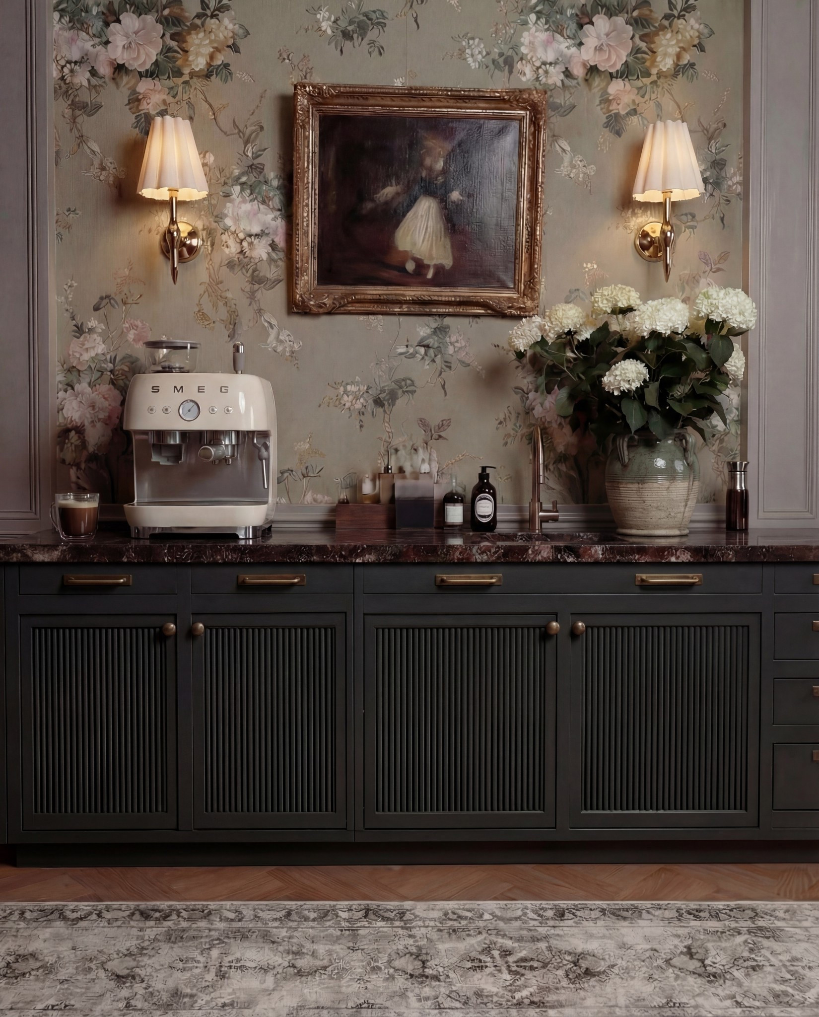This bar area feels moody, romantic, and old-world refined. Dark cabinetry grounds the space, while floral wallpaper and warm brass accents soften it with a lived-in, European charm. The styling feels curated rather than staged—perfect for slow mornings or evening drinks.

The marble countertop adds quiet luxury, layered with practical bar essentials and sculptural decor. Symmetrical wall sconces frame a vintage-style painting, creating a focal moment that feels intentional and timeless.

Shop the Look:
• Custom bar cabinetry (charcoal or deep green, reeded fronts)
• Brass cabinet pulls and knobs
• Dark marble or stone countertop (brown, burgundy, or black veining)
• Floral wallpaper (muted green, taupe, blush tones)
• Brass wall sconces with pleated fabric shades
• Framed vintage artwork (ornate gold frame)
• Smeg Espresso machine (cream or off-white)
• Ceramic mug or glass cup
• Wooden tray for bar essentials
• Glass bottles and barware
• Stoneware vase (aged or textured finish)
• White hydrangea floral arrangement
• Built-in sink with brass faucet
• Neutral vintage-style runner rug

#LTKHome #LTKSaleAlert #LTKFindsUnder100