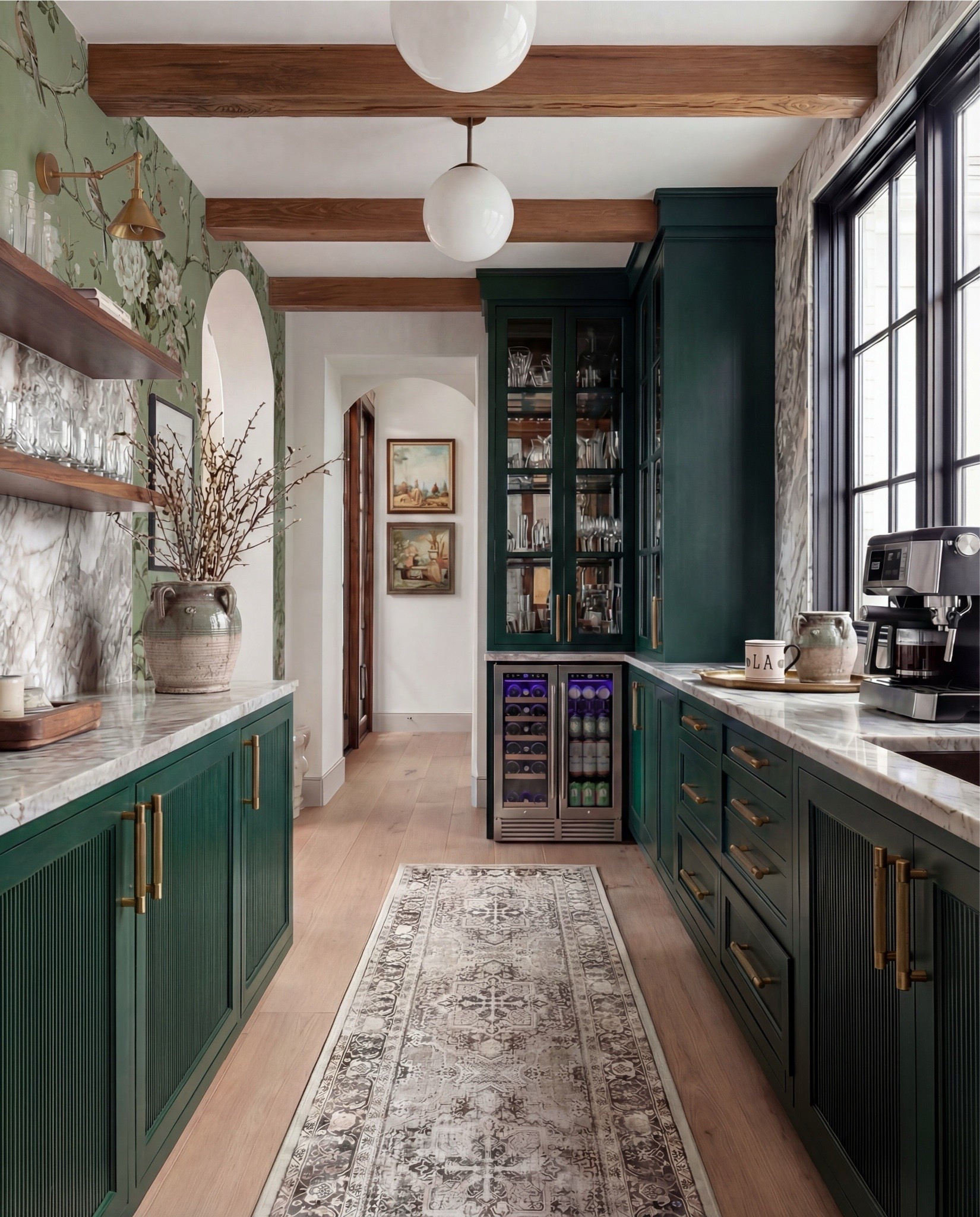 This cabinet moment is moody, refined, and quietly luxurious. The deep green cabinetry instantly grounds the space, while warm brass hardware and marble countertops keep it feeling elevated and timeless. Exposed wood beams add architectural warmth, and the glass-front uppers make everyday glassware feel like decor.

The styling is subtle but intentional—ceramic vessels, open shelving with clear stemware, and a vintage-style runner that softens the galley layout without competing with the finishes.

Shop the Look:
• Deep green paneled cabinetry
• Brass cabinet pulls + knobs
• Marble countertops (soft gray veining)
• Glass-front upper cabinets
• Built-in beverage/wine fridge
• Vintage-style runner rug (neutral tones)
• White globe pendant lights
• Exposed wood ceiling beams
• Open wood shelving
• Clear glassware set
• Ceramic vase with branches
• Countertop coffee machine
• Stoneware mugs + tray
• Light wood flooring
• Arched doorway with framed artwork
• coffee machine
• brass gold faucet 
• green vase
• letter bistro cup 
• globe pendant light 
Brass wall sconce 
Brass hand pulls

#LTKSaleAlert #LTKHome