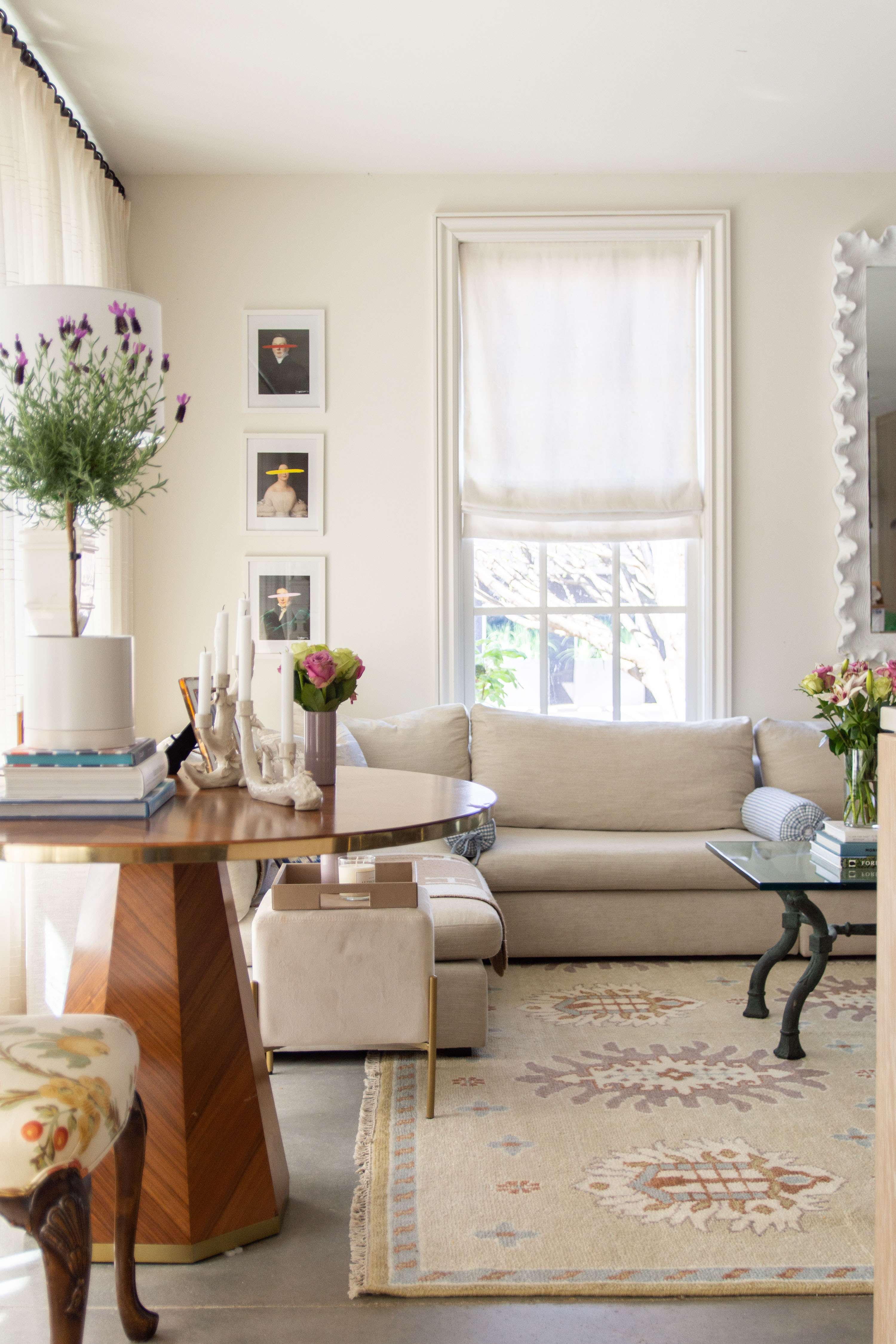 Bright, cozy, and effortlessly chic living room design featuring a neutral linen sofa, layered with soft roll pillows for a timeless look. The space is anchored by a vintage-inspired Turkish rug in warm beige tones with subtle pops of blue and rust, adding texture and character. A round wood pedestal table styled with stacked design books, taper candles, and fresh florals creates the perfect mix of modern and classic decor. White Roman shades filter natural light for an airy feel, while the gallery wall of contemporary art adds a sophisticated touch. Finished with a sculptural glass coffee table and fresh greenery, this living room blends modern elegance with comfortable charm. 

 