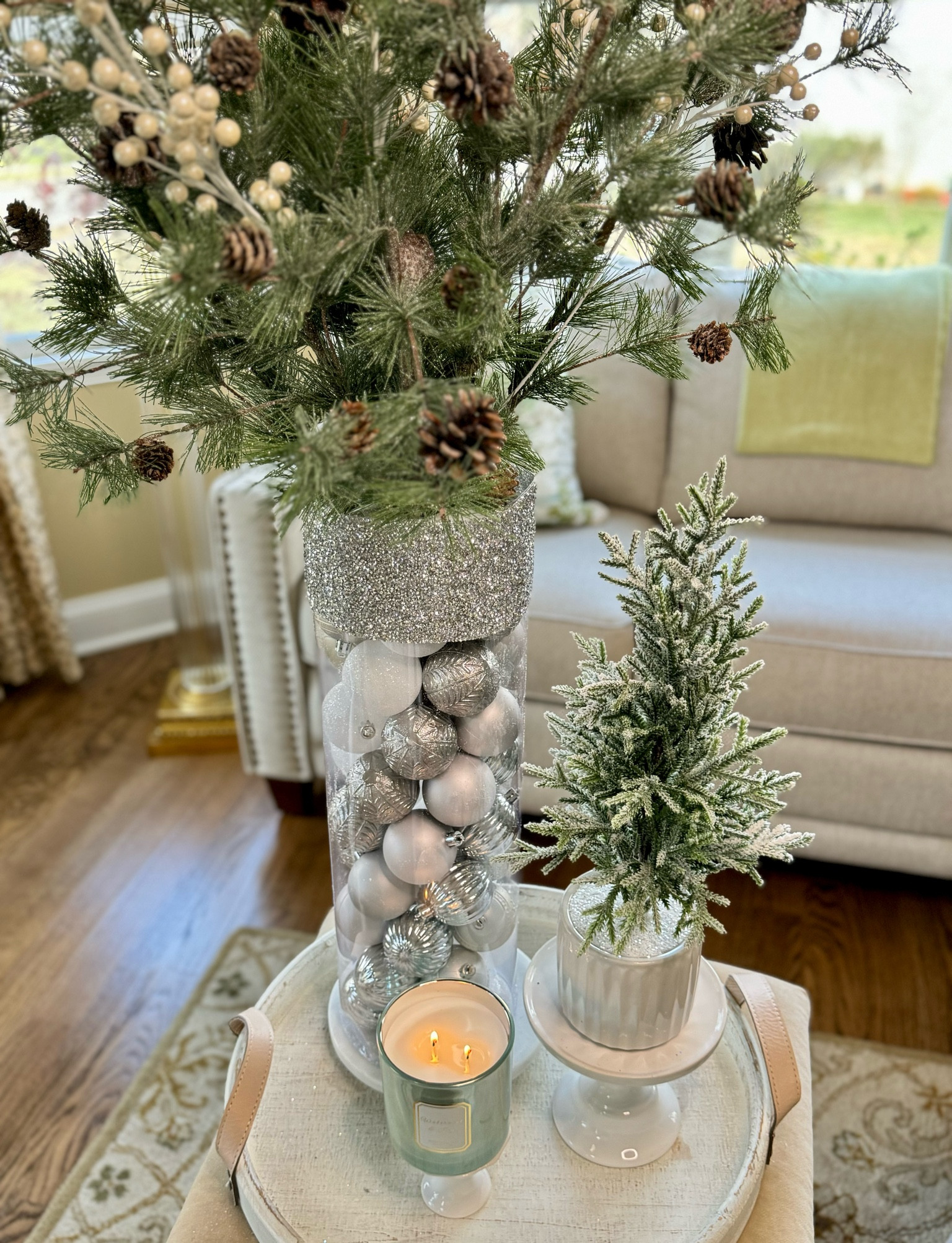 Elegant winter holiday decor featuring a sparkling vase filled with silver Christmas ornaments, frosted pine branches, and pinecones. The cozy setup is completed with a festive candle and a miniature frosted Christmas tree, perfect for creating a warm and inviting seasonal ambiance. 

#LTKHoliday #LTKStyleTip #LTKHome