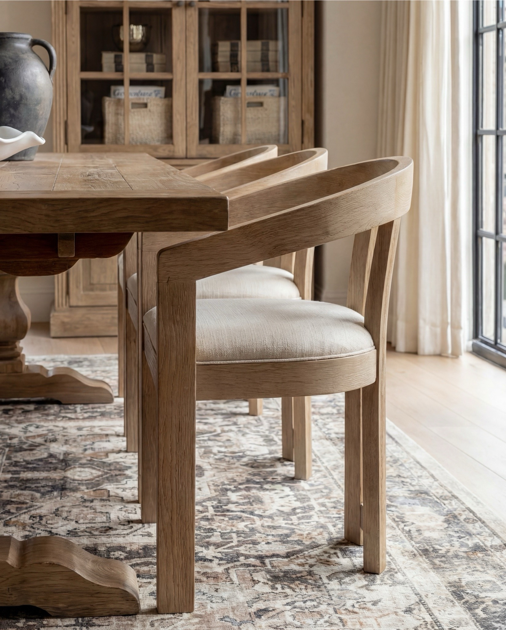 Shop the Look:
• Solid wood pedestal dining table (warm oak tone)
• Light wood dining chairs with upholstered seats
• Neutral vintage-style area rug (soft brown pattern)
• Brass chandelier with tapered fabric shades
• Exposed wood ceiling beams
• Large wood and glass display cabinet
• Fluted wood sideboard cabinet with brass hardware
• Black ceramic table lamp with linen shade
• Framed neutral textile wall art
• Matte black decorative vase
• Wood bead garland with tassels
• Decorative wood tray with ceramic bowl
• Small abstract sculpture decor
• Olive tree in stone planter
• White scalloped ceramic centerpiece bowl
• Dark ceramic pitcher vase (table decor)
• Glass hurricane candle holder
• Sheer neutral curtains

#LTKHome #LTKSaleAlert